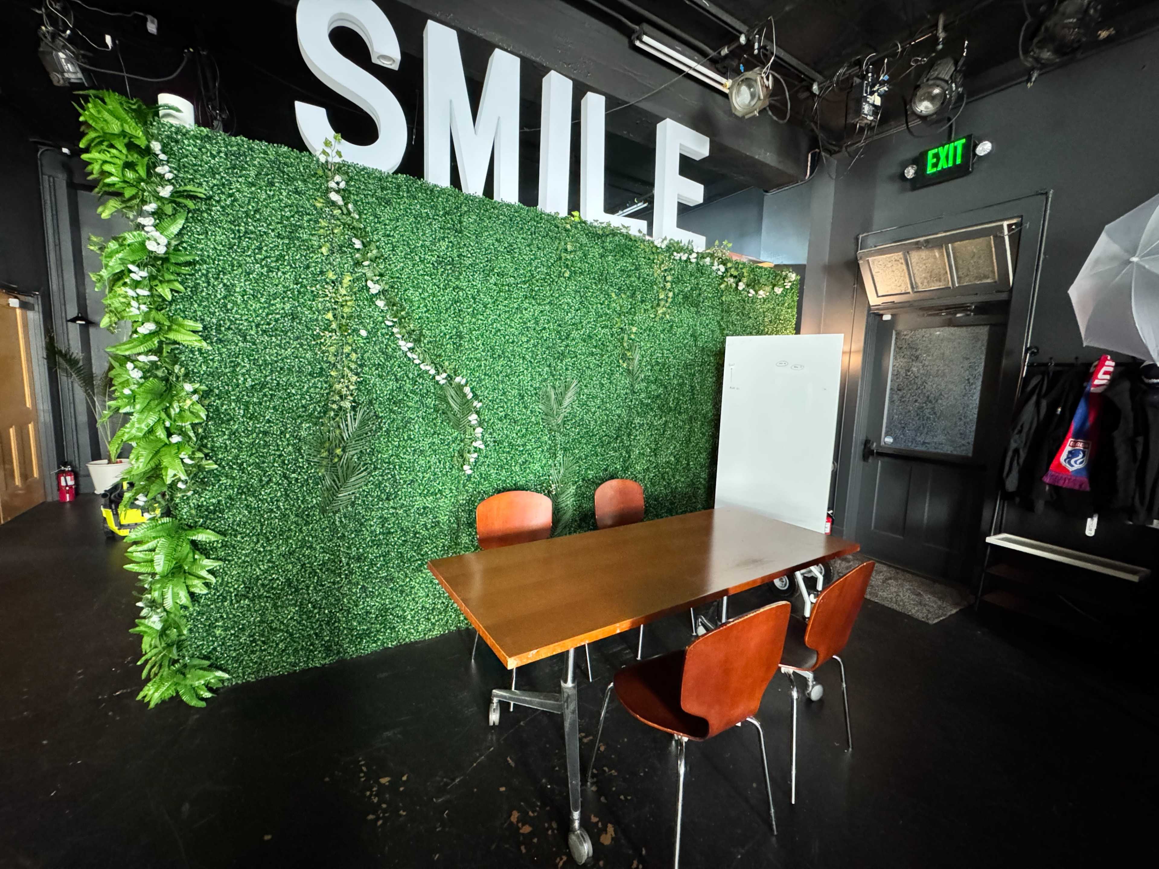 The image shows a small meeting space featuring a wooden table surrounded by four chairs, with a greenery-covered wall and the word "SMILE" displayed above.
