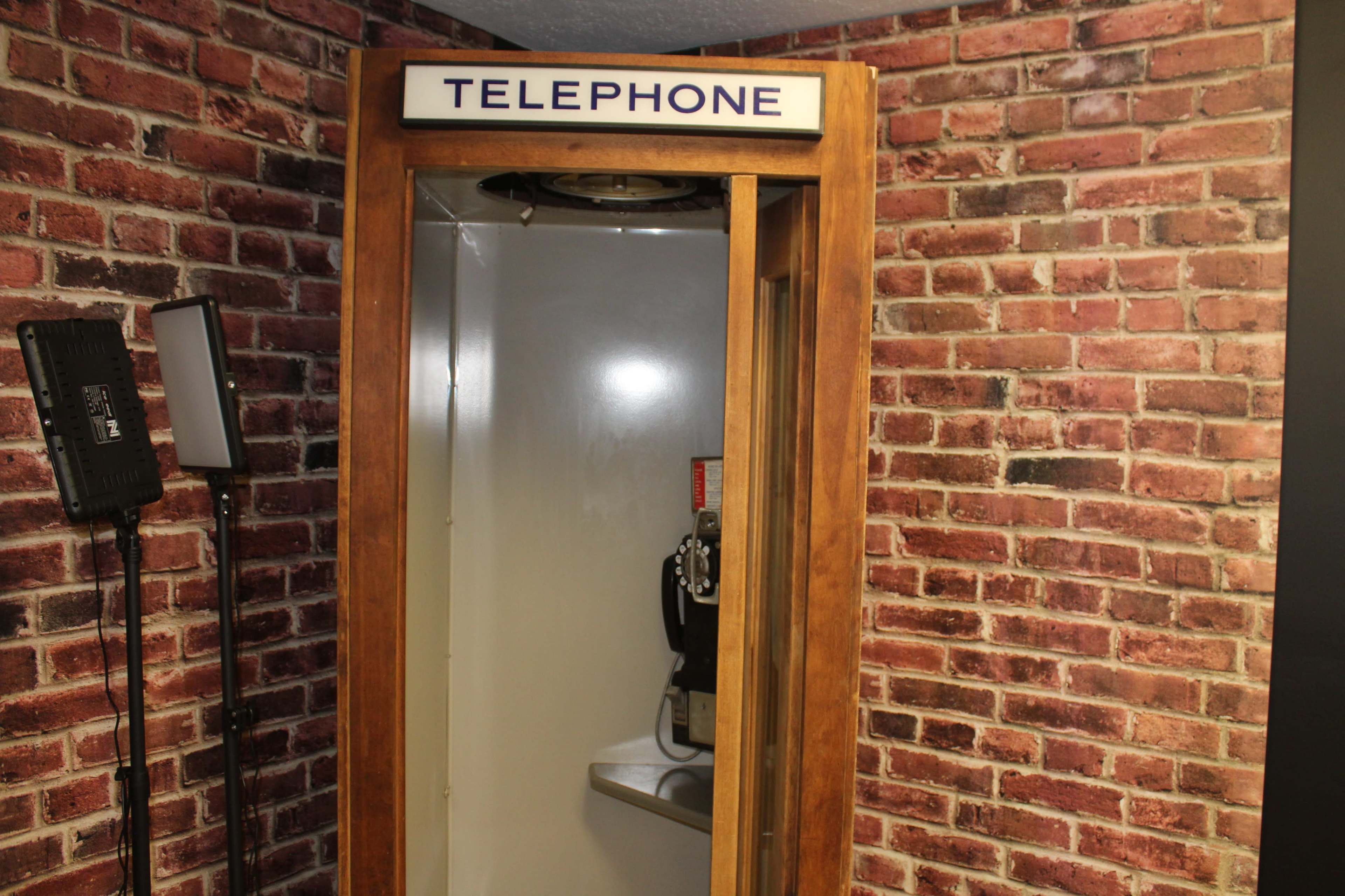 The image shows a wooden phone booth with the word "TELEPHONE" displayed on the top, set against a brick wall background.