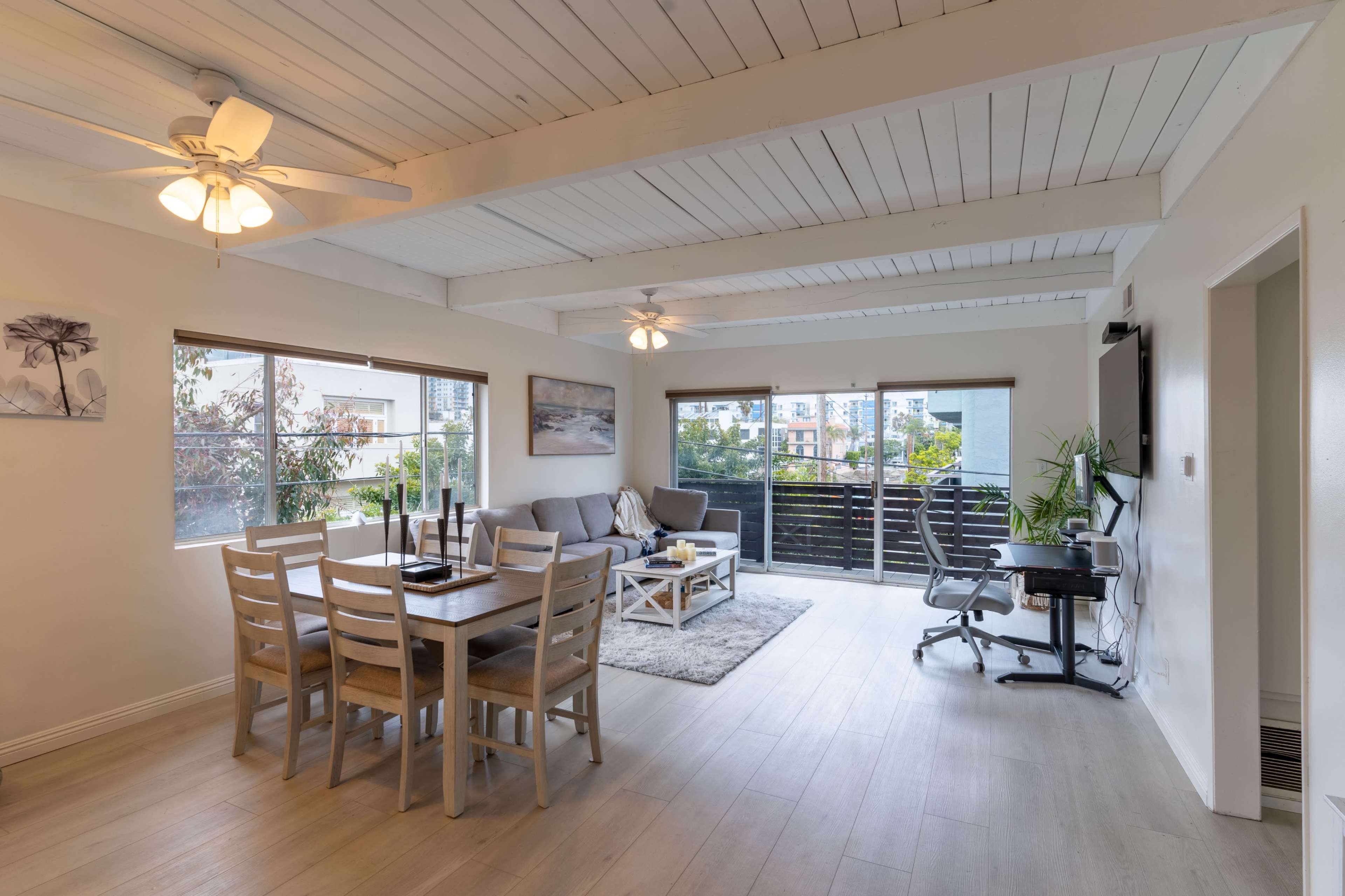 The image shows a modern living and dining area with a light wood dining table, a comfortable seating arrangement, and large windows overlooking a balcony.