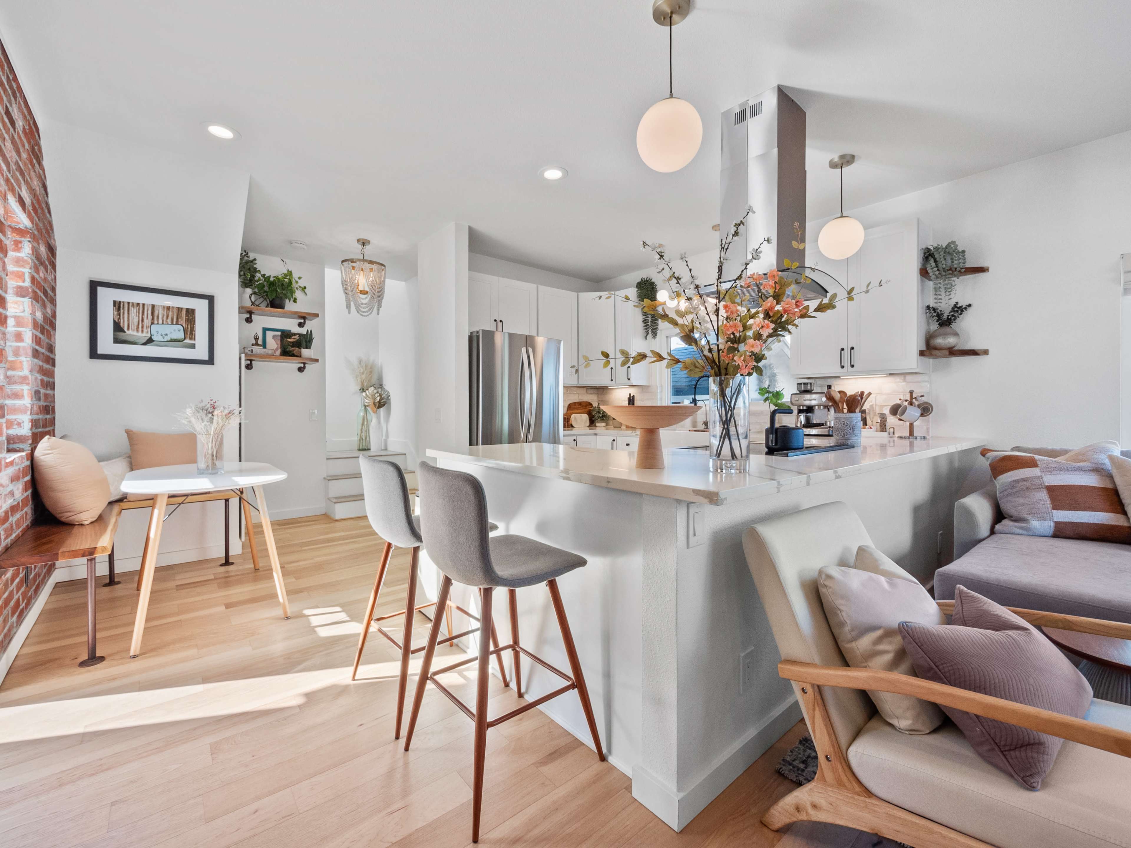 A modern kitchen and dining area with a central island, bar stools, and a small dining table beside a brick wall.