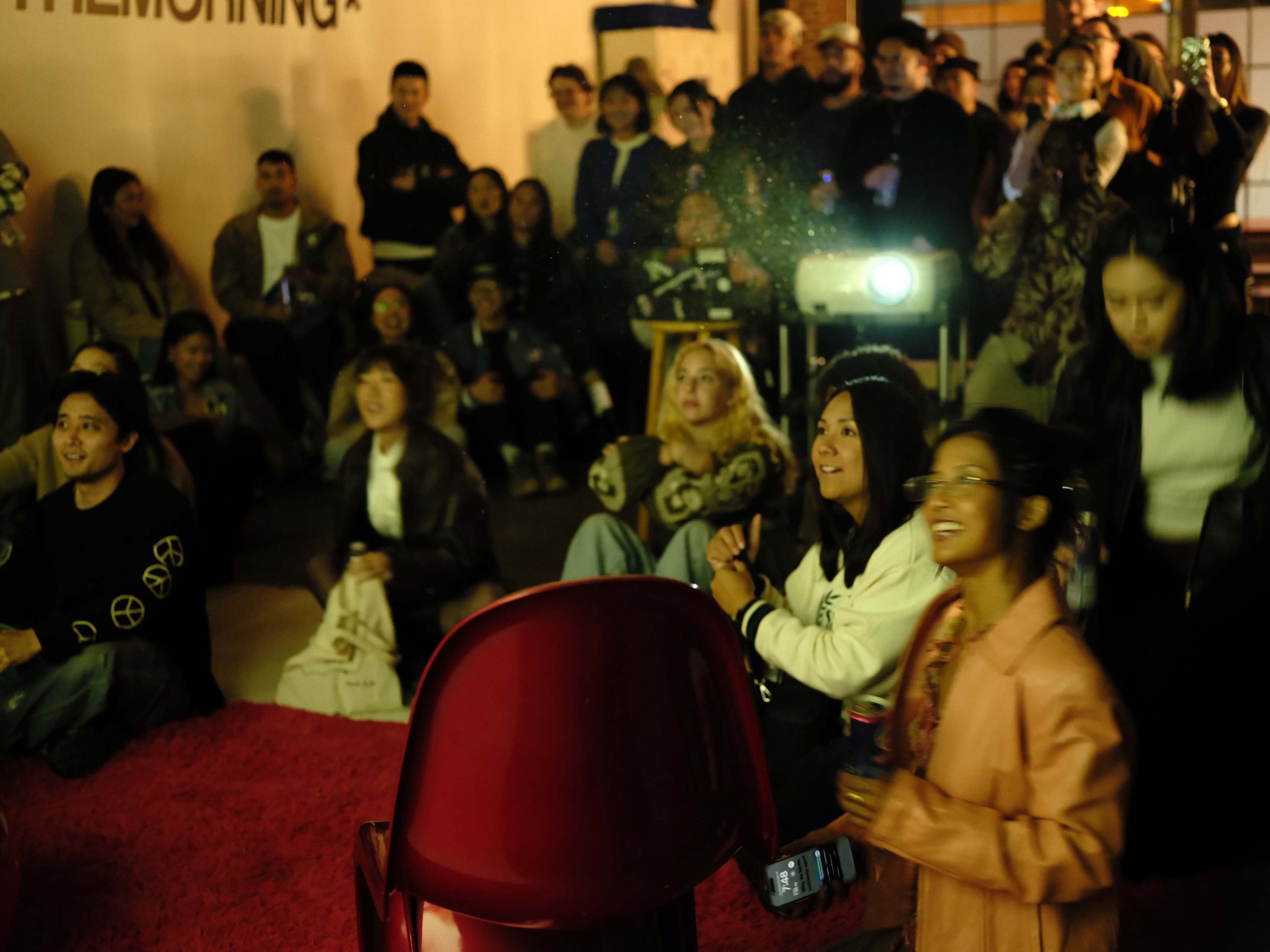 A crowd of people is gathered indoors, watching a presentation projected on a screen while seated on the floor and chairs.