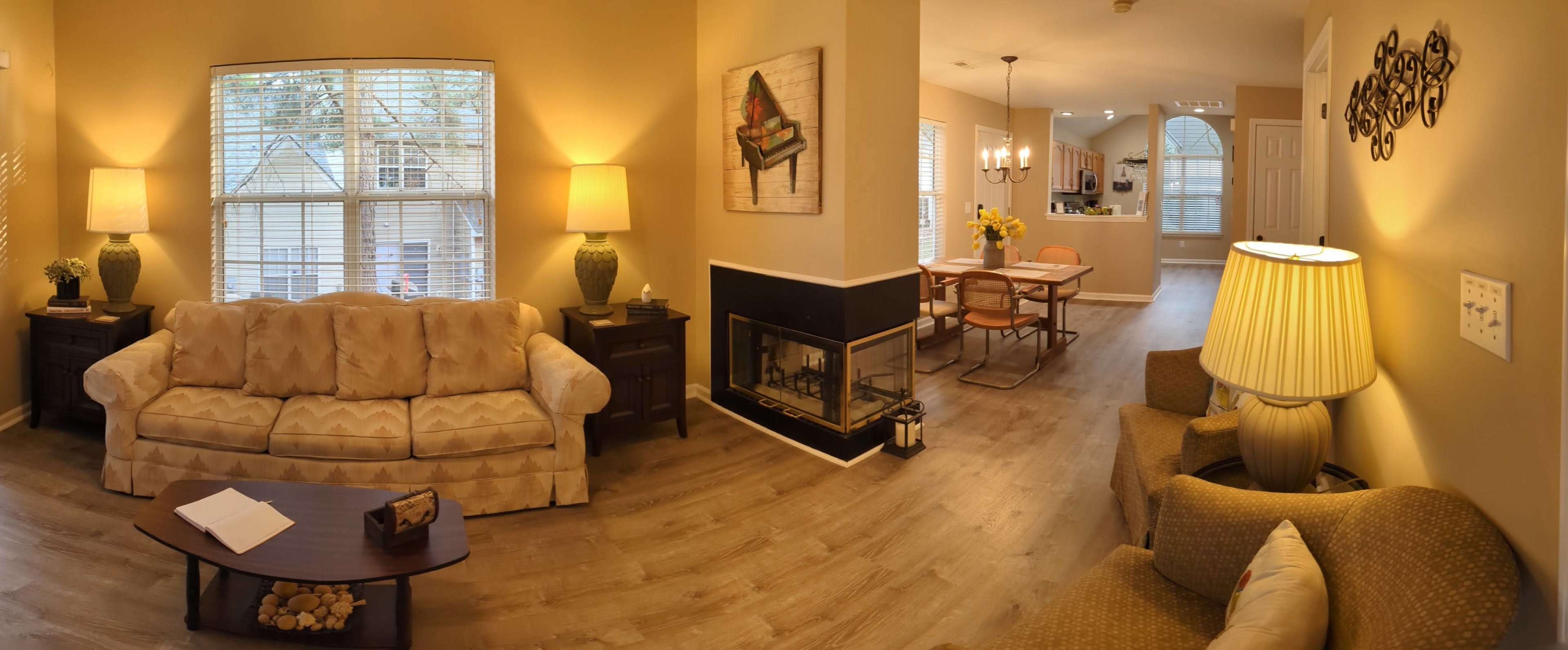 A well-lit living room featuring a beige couch, a cozy armchair, a coffee table, and a fireplace, with a view into a dining area and kitchen beyond.
