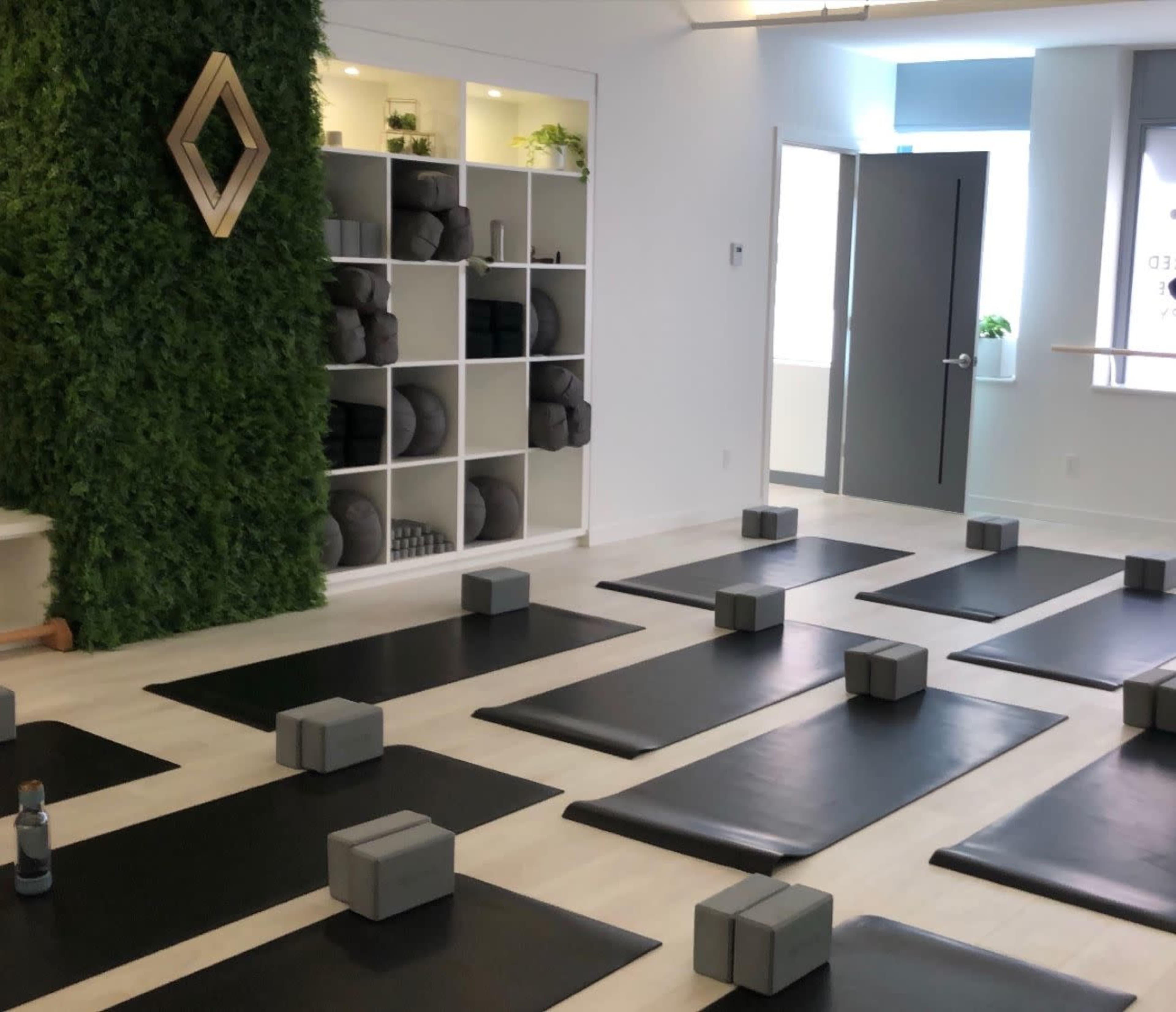 A yoga studio with several black mats arranged in rows, gray yoga blocks placed on each mat, and a green wall featuring shelves stocked with yoga accessories.