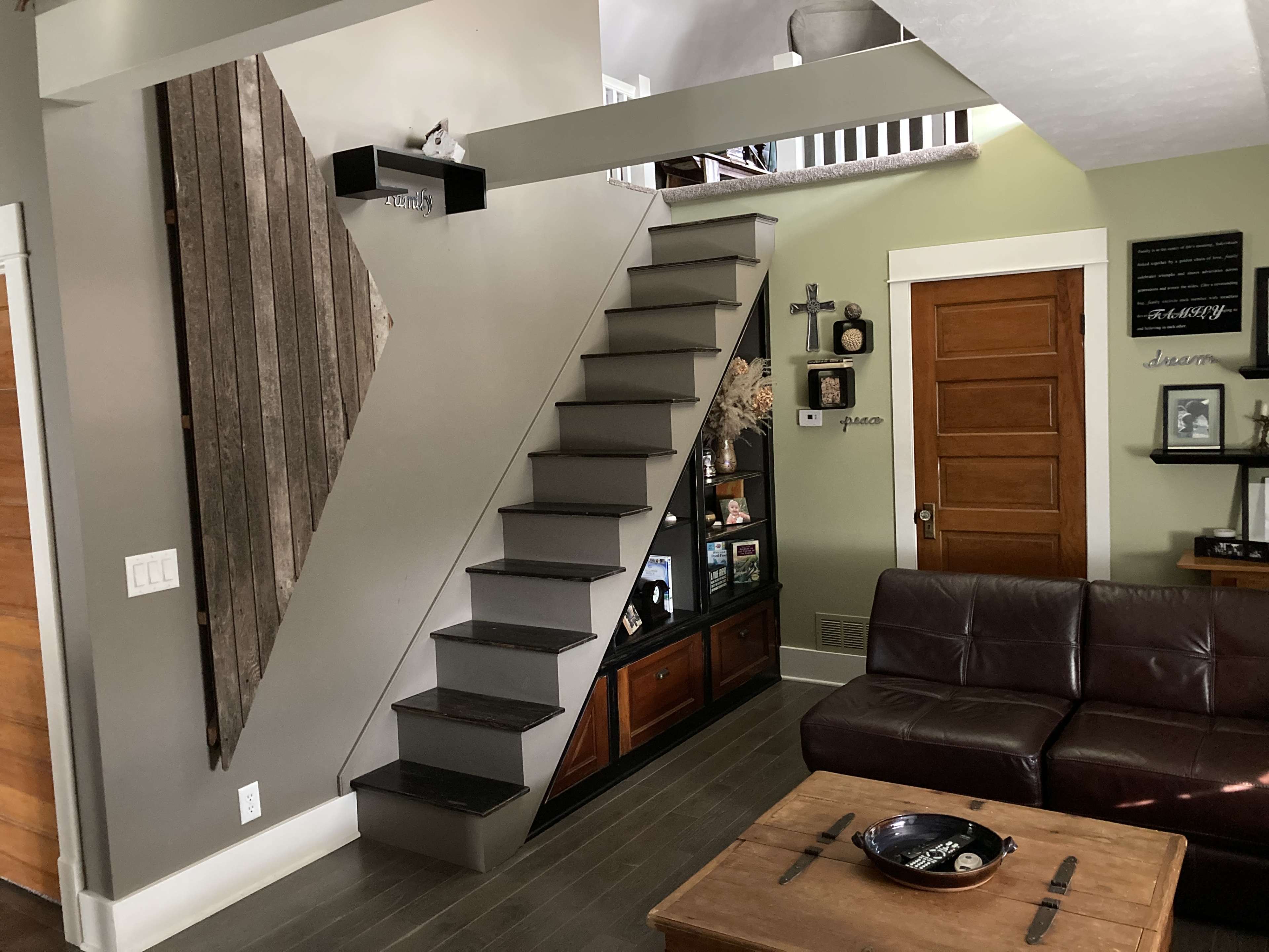 The image shows a modern living room with a staircase made of metal steps leading to an upper level, alongside a wooden wall decoration and a brown leather sofa.