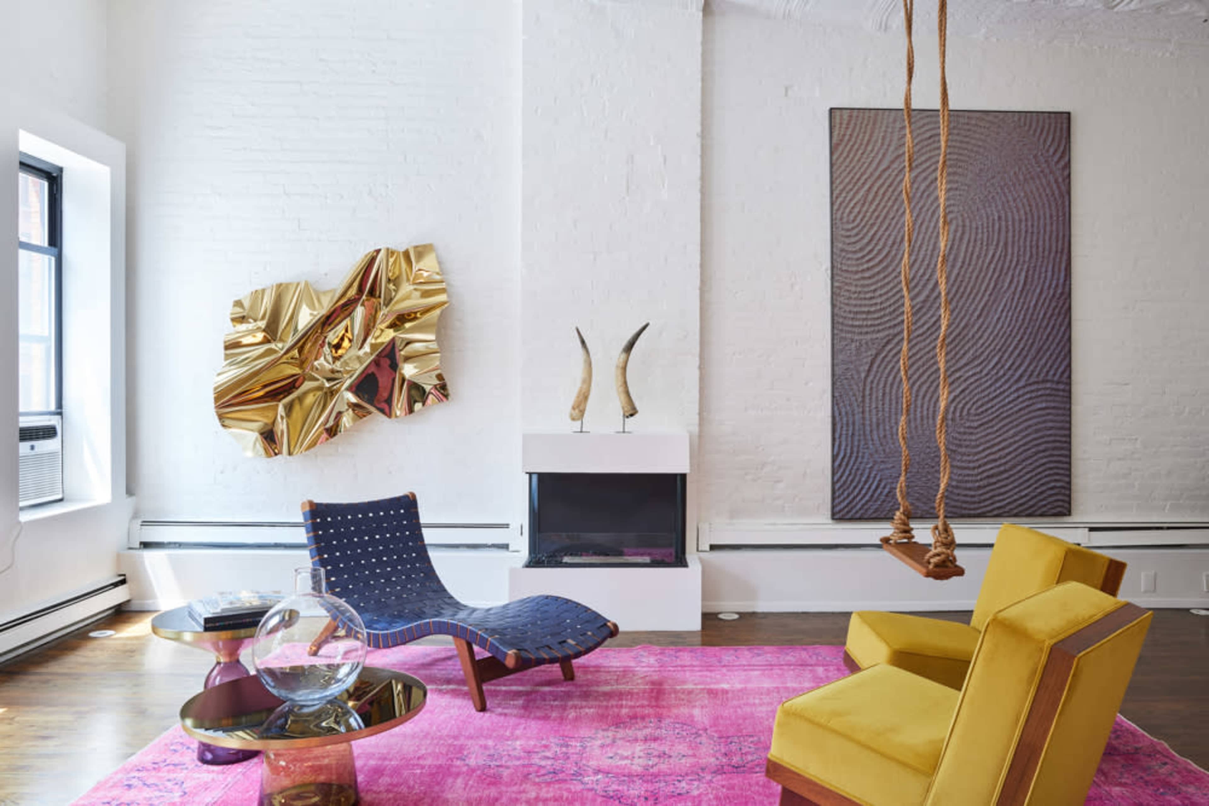 A modern living room featuring a blue chair, a yellow armchair, a pink rug, a glass coffee table, a white fireplace, and a large wall art piece.
