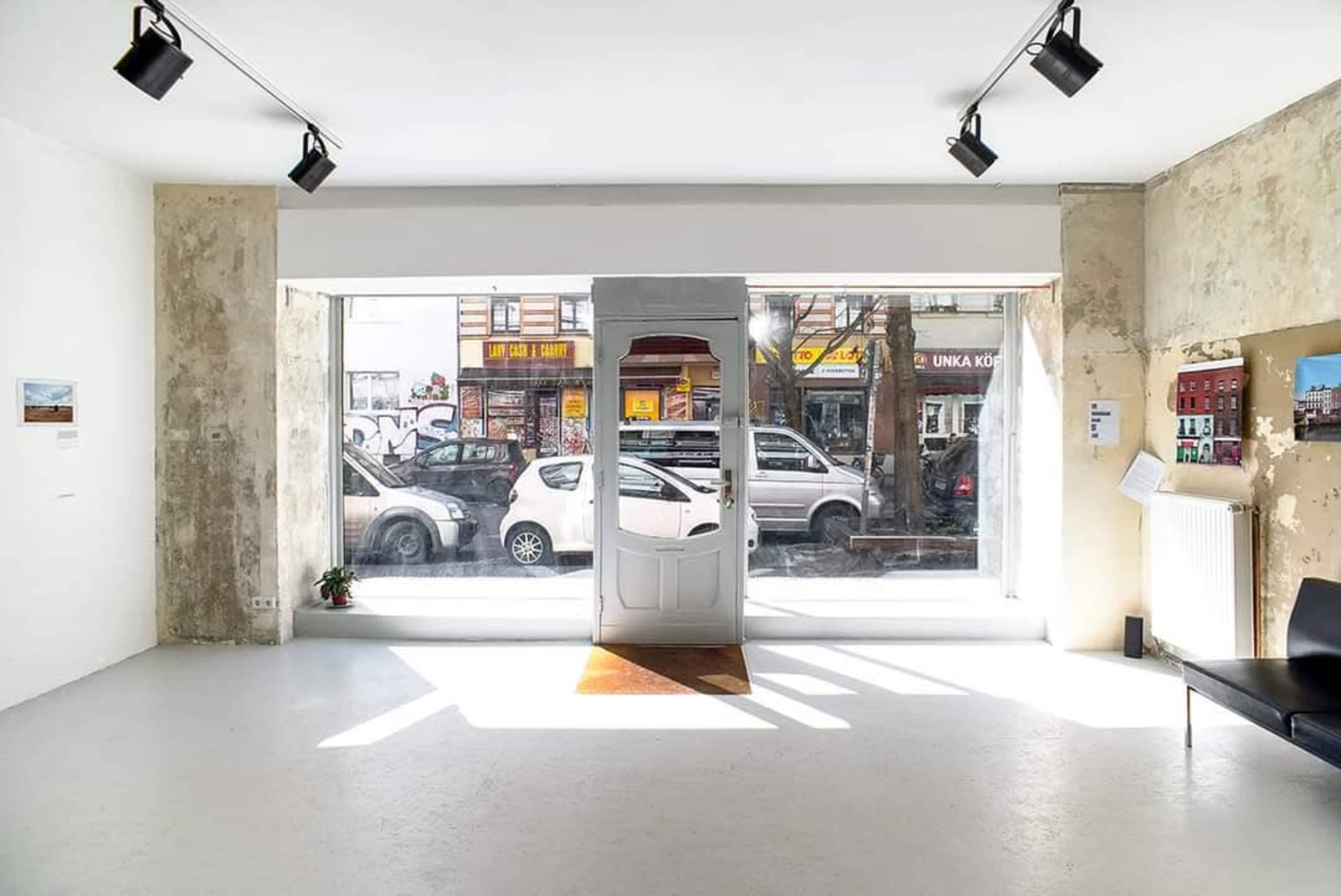 The image shows a minimalist interior of an art gallery with a large window displaying cars and buildings outside.