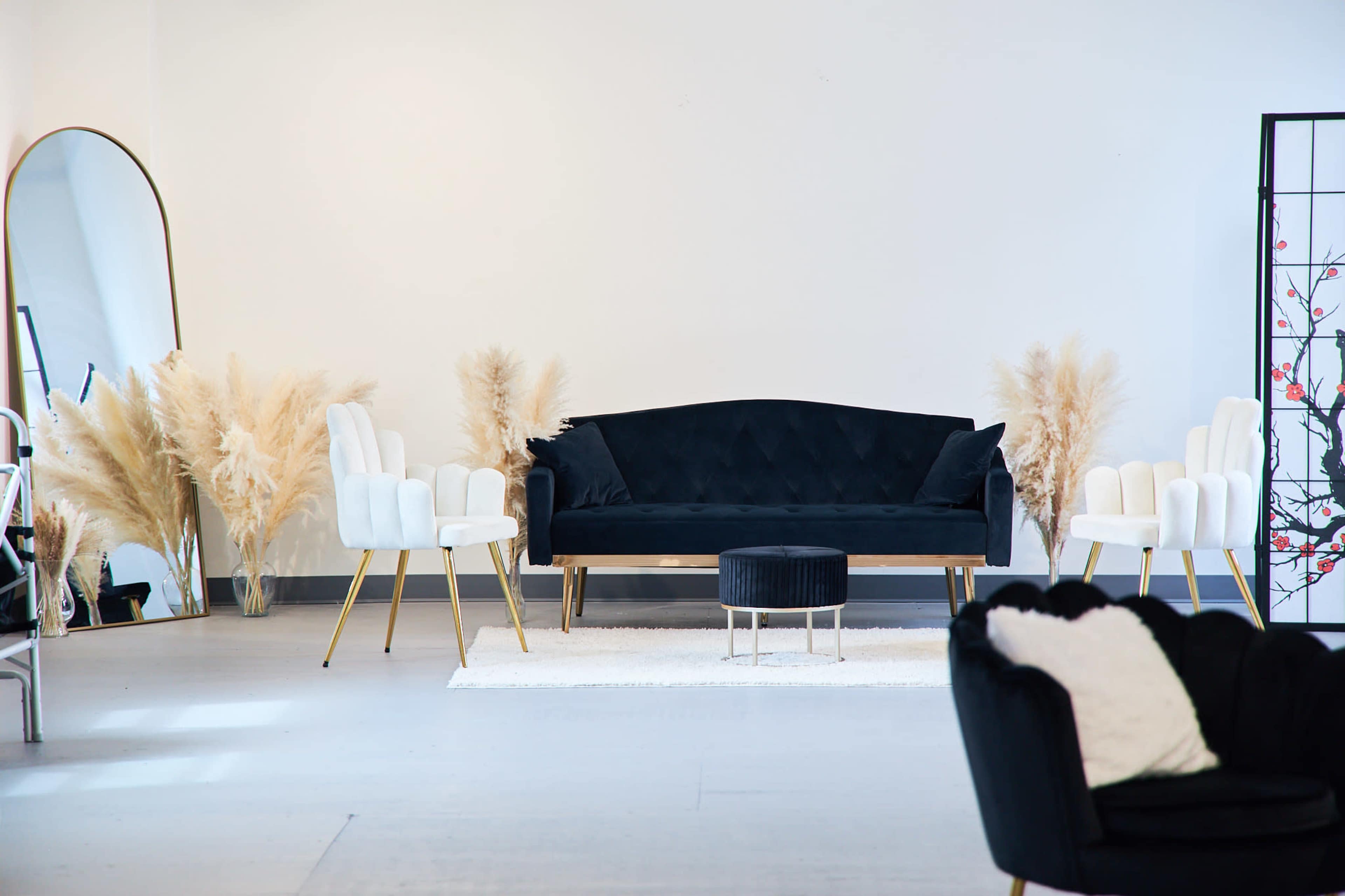 A modern lounge area with a black sofa, two white chairs, a small round table, pampas grass decor, and a large mirror against a white wall.