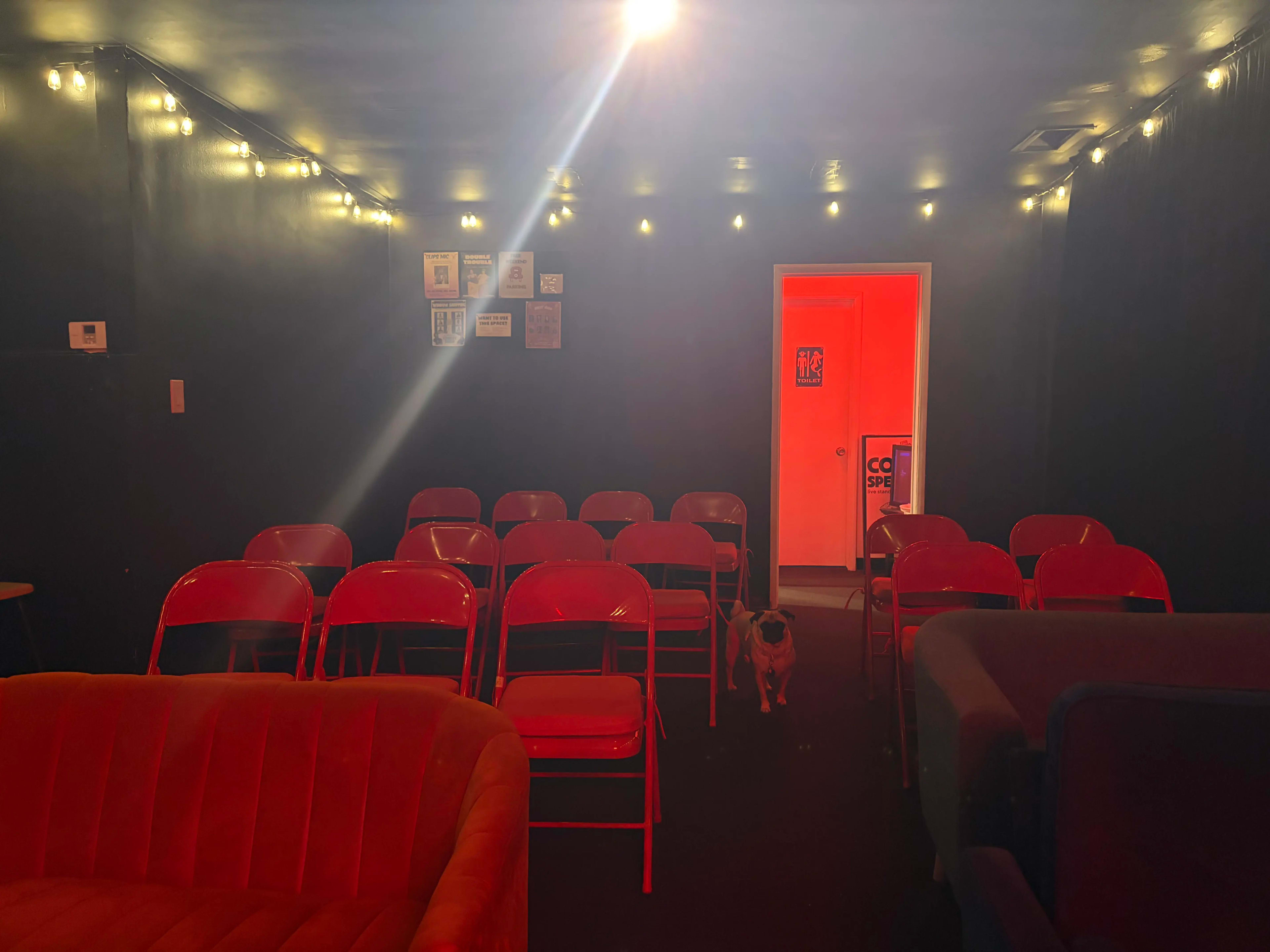 A small theater room with red chairs arranged in rows, dim lighting, and a door emitting orange light at the back.
