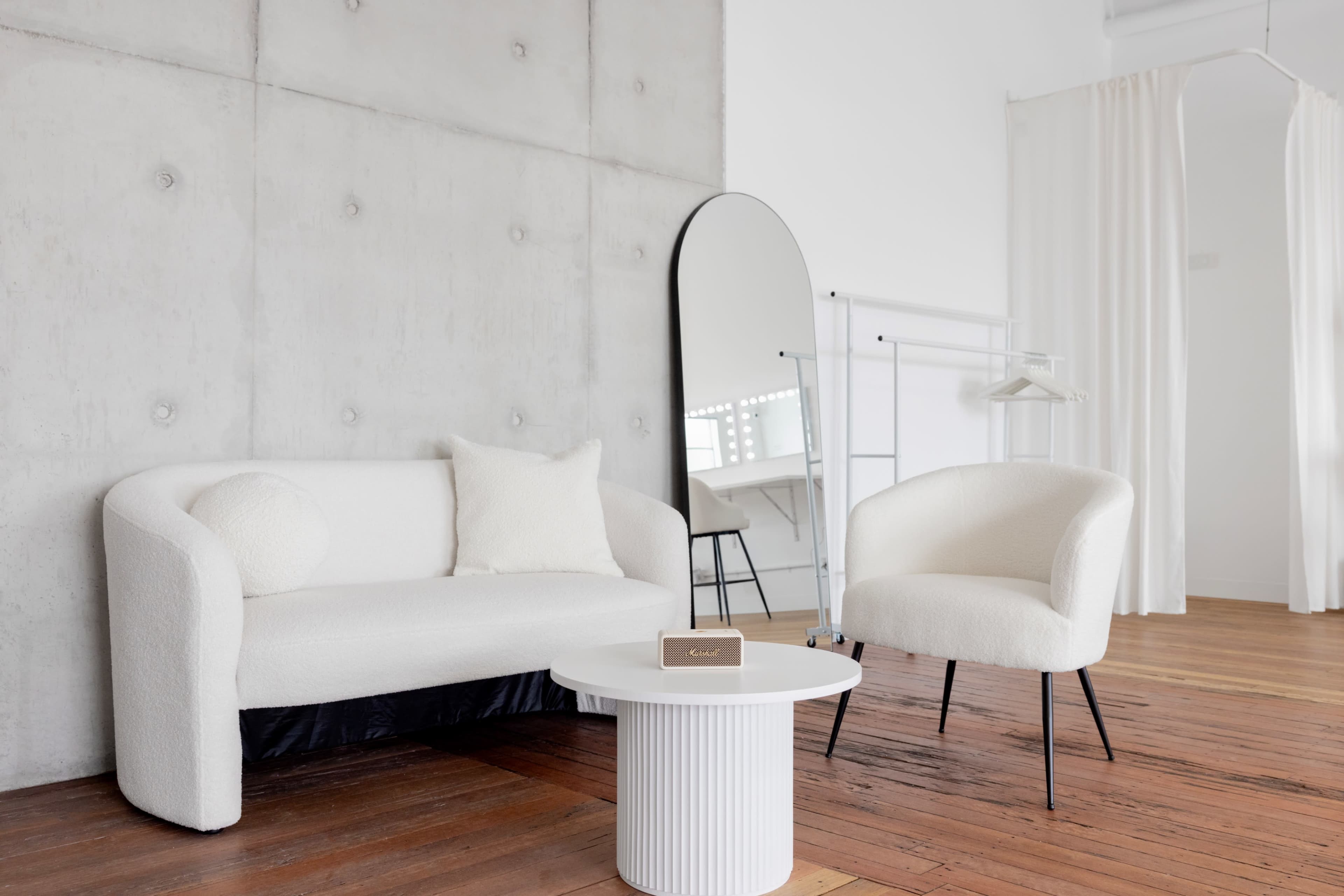 A modern interior with a white sofa, a round white table, and a large mirror against a concrete wall, complemented by wooden flooring.