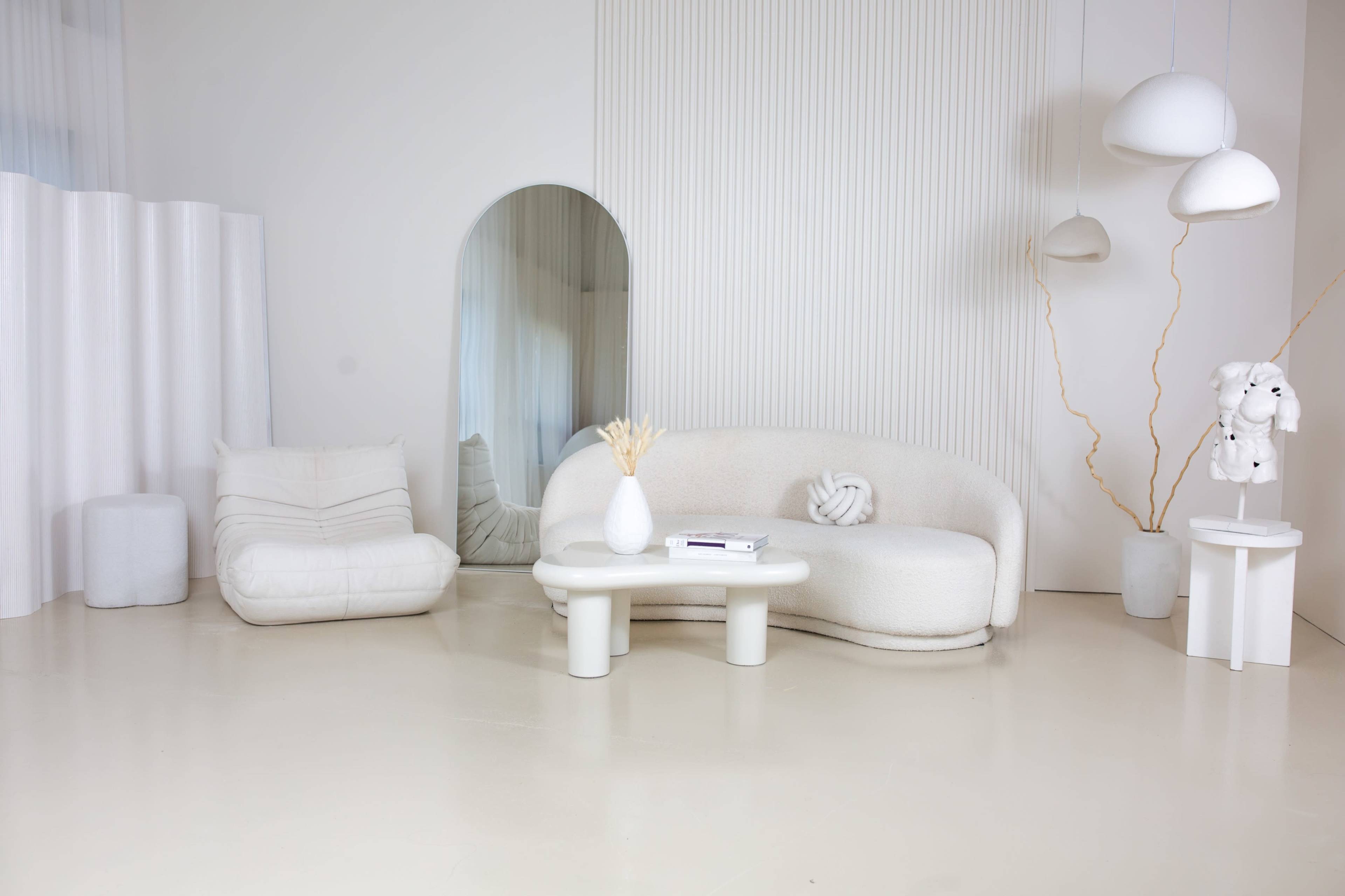 The image features a minimalist interior design with white furniture, including a curved sofa, a simple table, and an accent chair, all set against a light-colored backdrop with various shapes and textures.