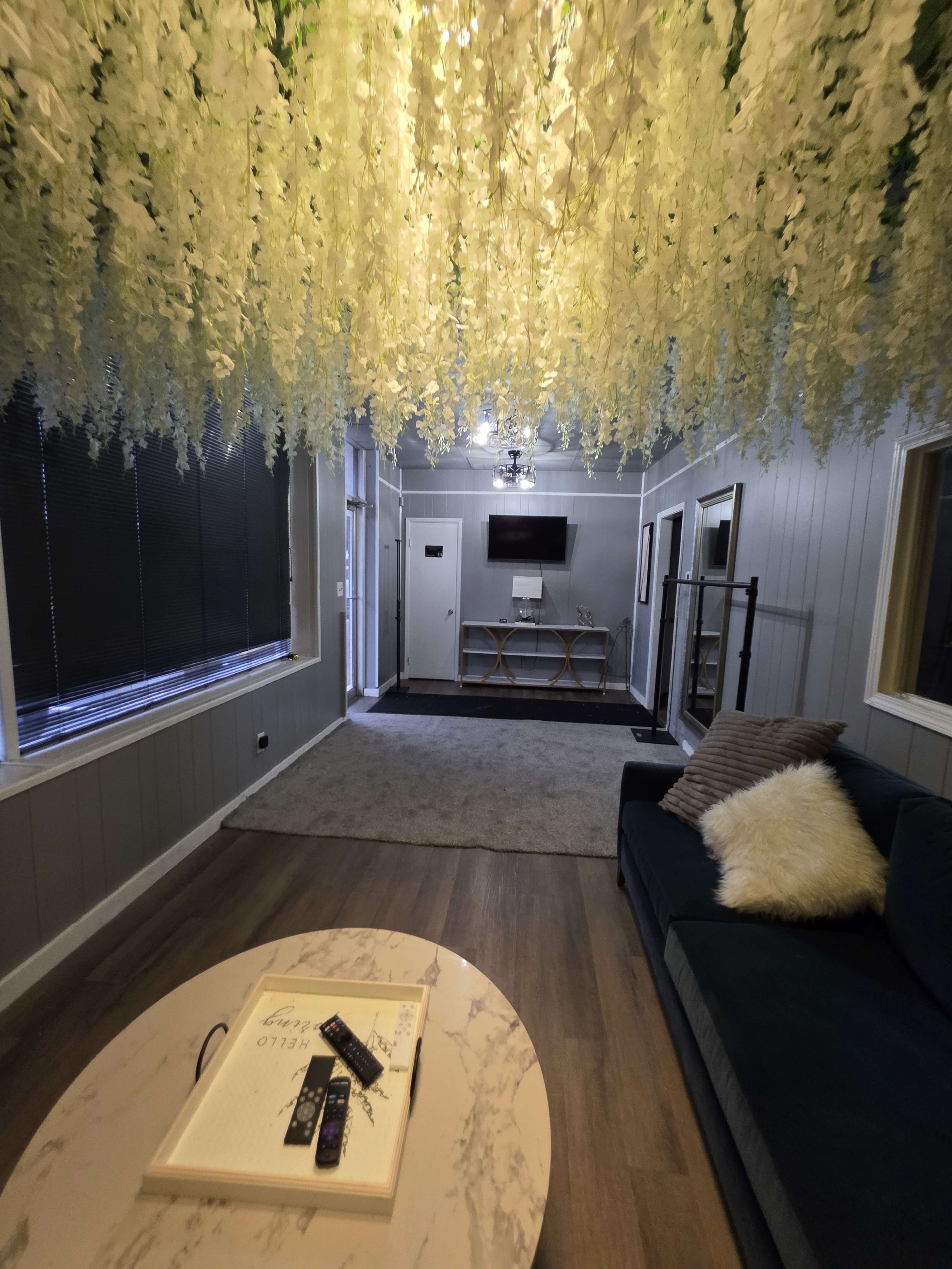 The image shows a living room with a dark sofa, a coffee table featuring a remote, and a ceiling adorned with hanging artificial flowers.