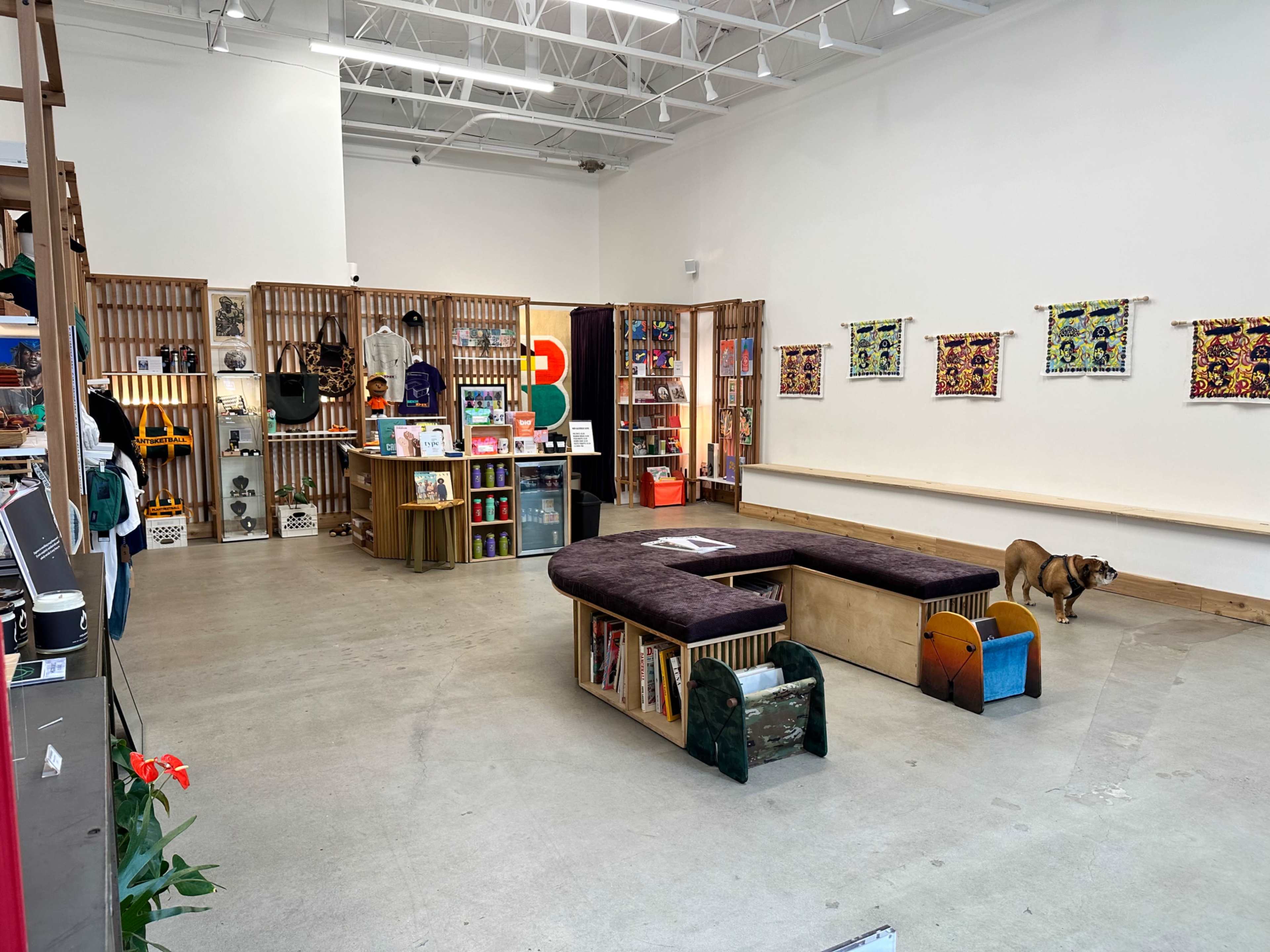 The image shows an interior of a retail space with a central seating area, shelves displaying various products, and a small dog on the floor.
