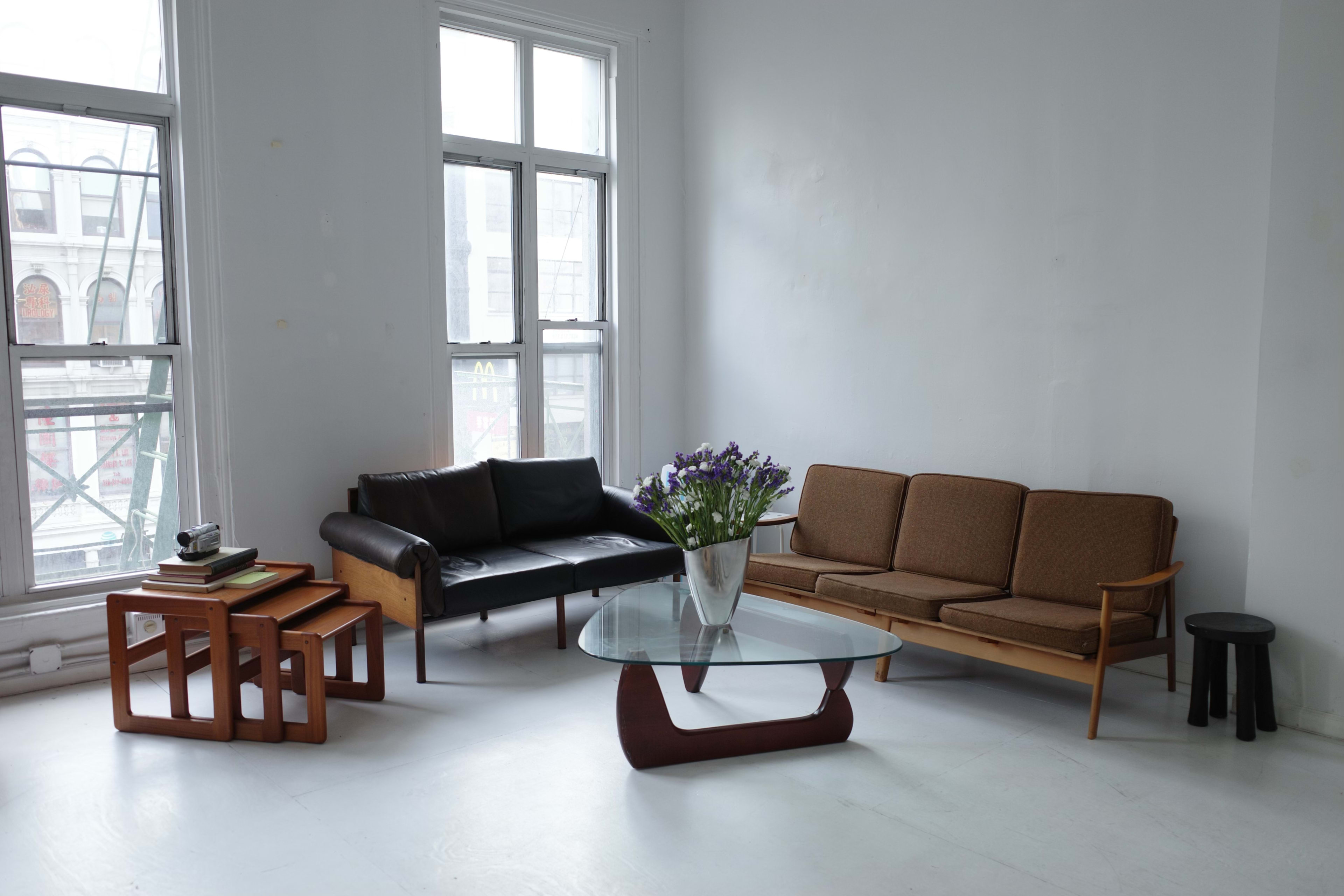 A modern living room features a black leather sofa, a brown upholstered couch, a glass coffee table, wooden side tables, and a vase of flowers, all set against white walls and large windows.