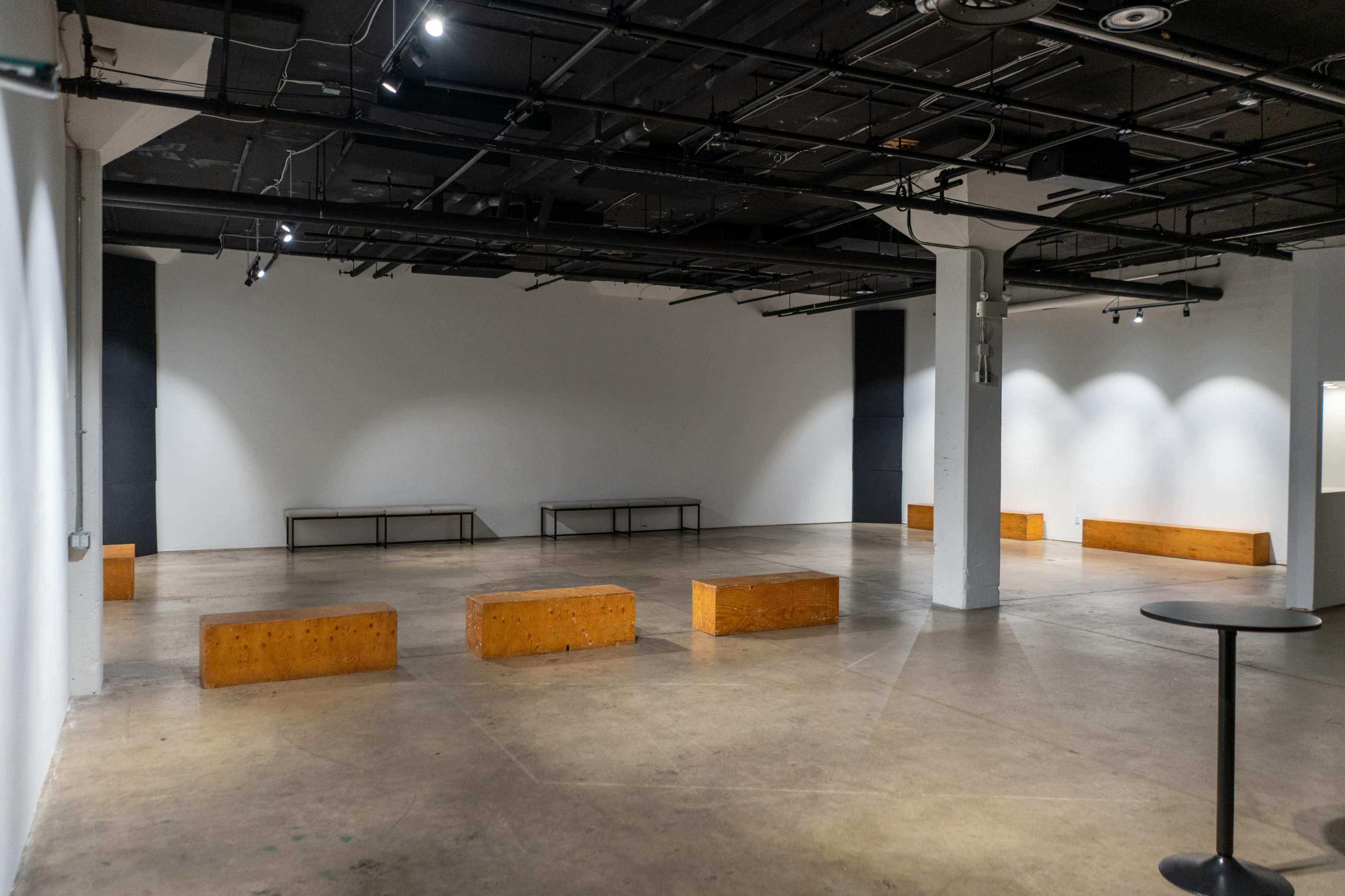 The image shows an empty indoor space with concrete flooring, minimalist wooden benches, and overhead lighting.