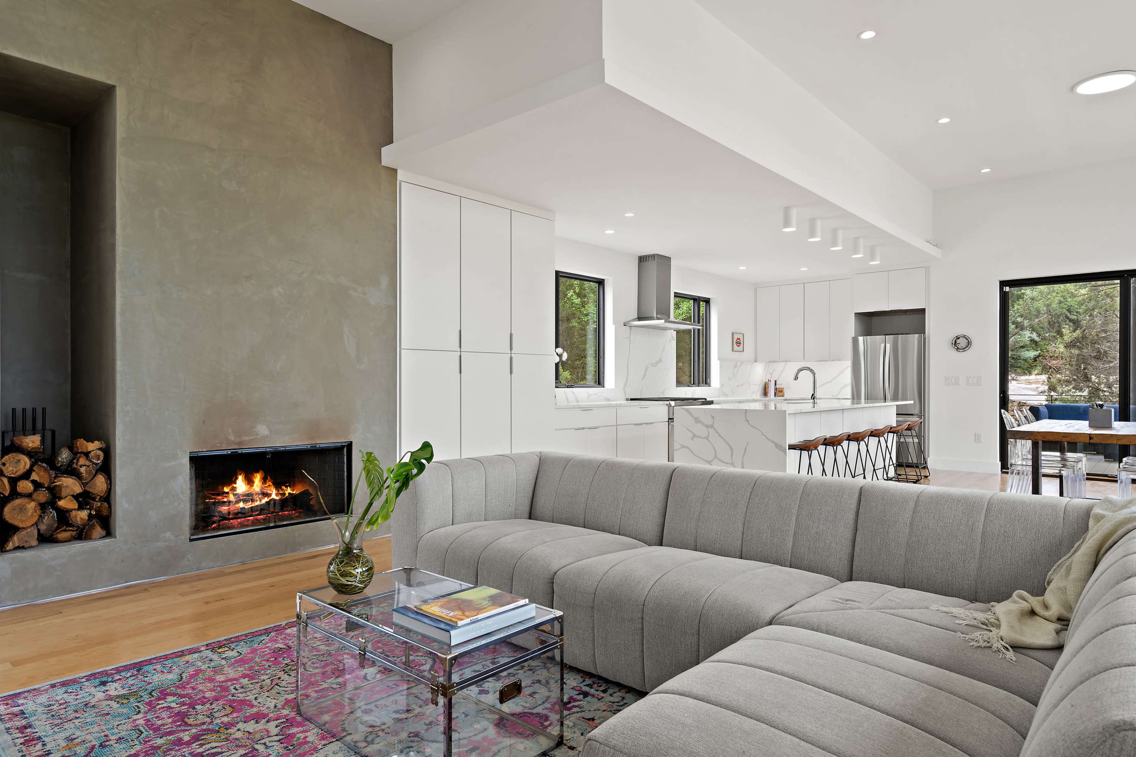 A modern living space features a gray sectional sofa, a glass coffee table, a wood-burning fireplace, and a view of an open kitchen.