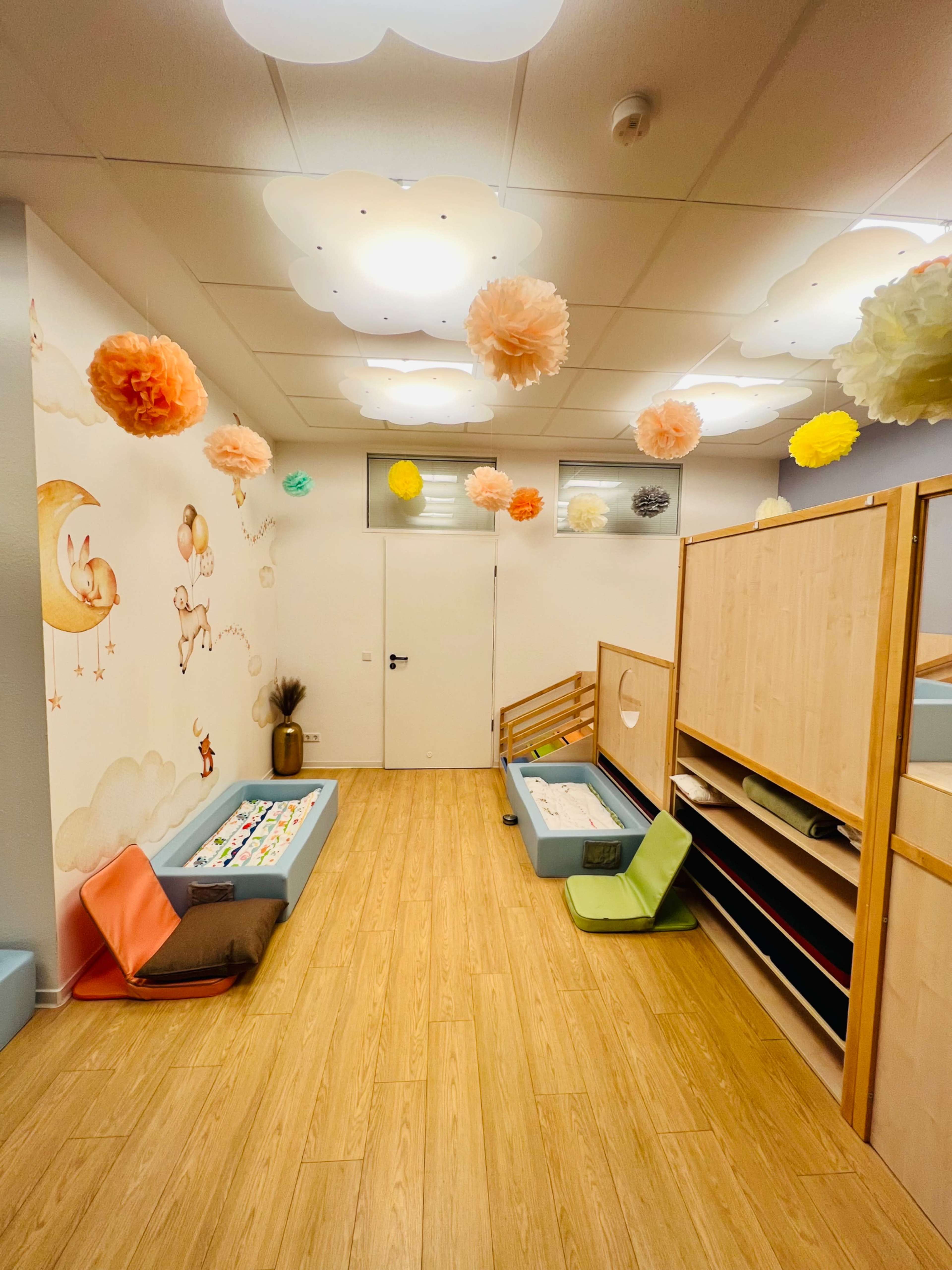 The image shows a brightly decorated children's playroom featuring colorful paper decorations hanging from the ceiling, a wall mural with clouds and a moon, and cozy seating areas with benches.