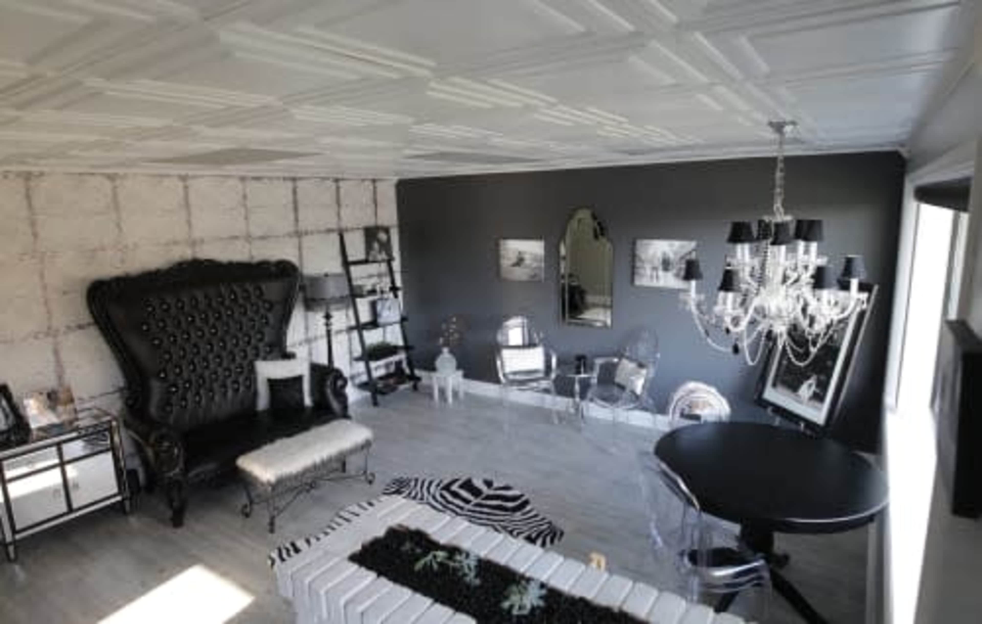 A modern living room featuring a large black tufted armchair, a mirrored shelf, clear acrylic chairs, a round black dining table, and a decorative chandelier against a textured wall.
