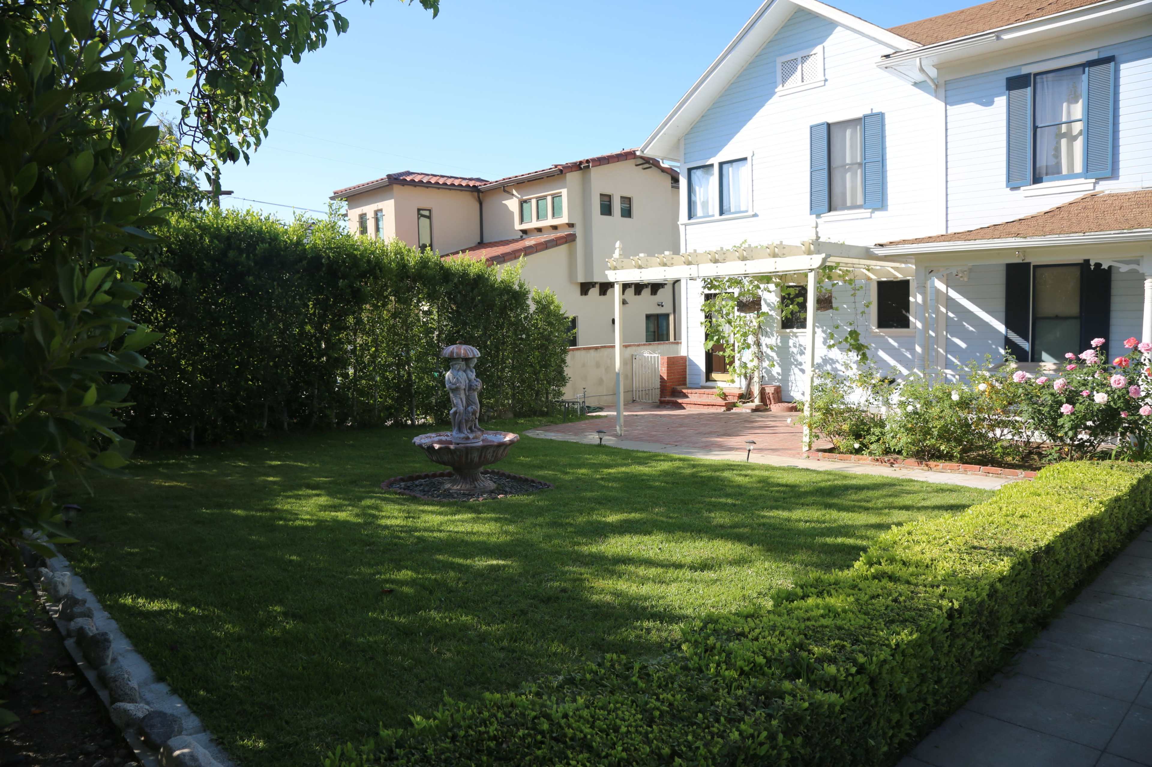 A well-maintained garden with a fountain centered in a lawn, surrounded by hedges and featuring a house with a porch in the background.