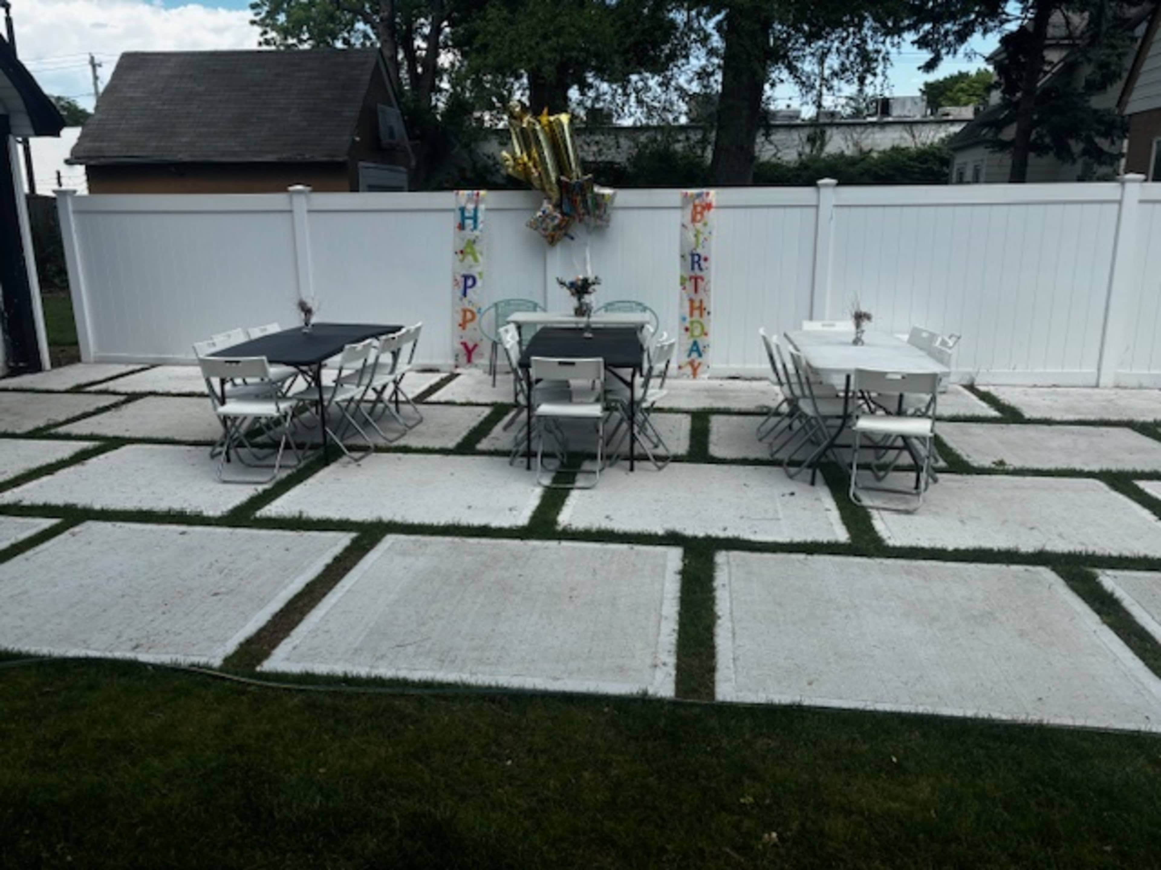 A birthday celebration setup features two tables with chairs arranged on a grassy patio adjacent to a white fence decorated with colorful birthday banners.