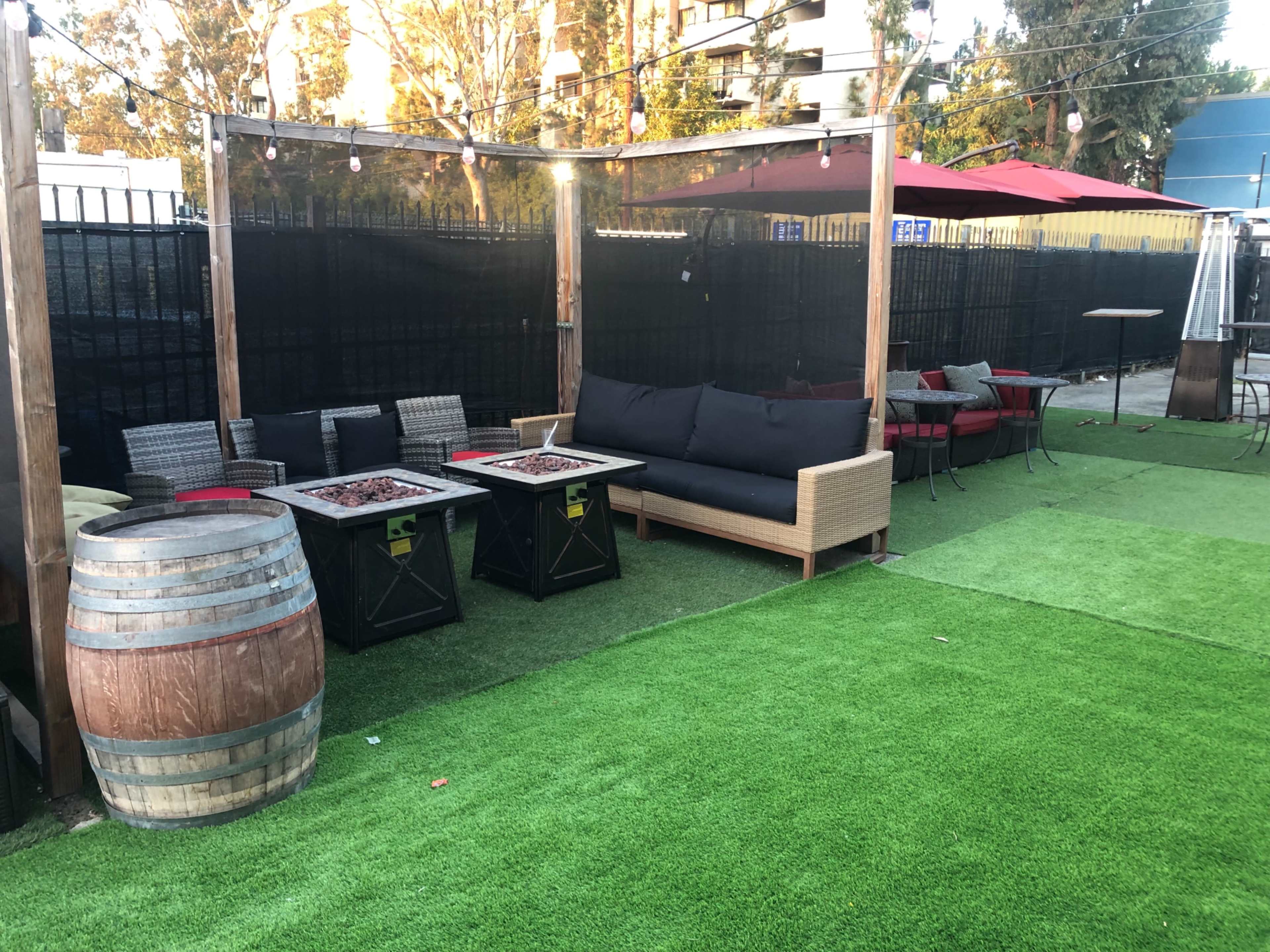 The scene features an outdoor seating area with synthetic grass, several tables, comfortable couches, and a wooden barrel.