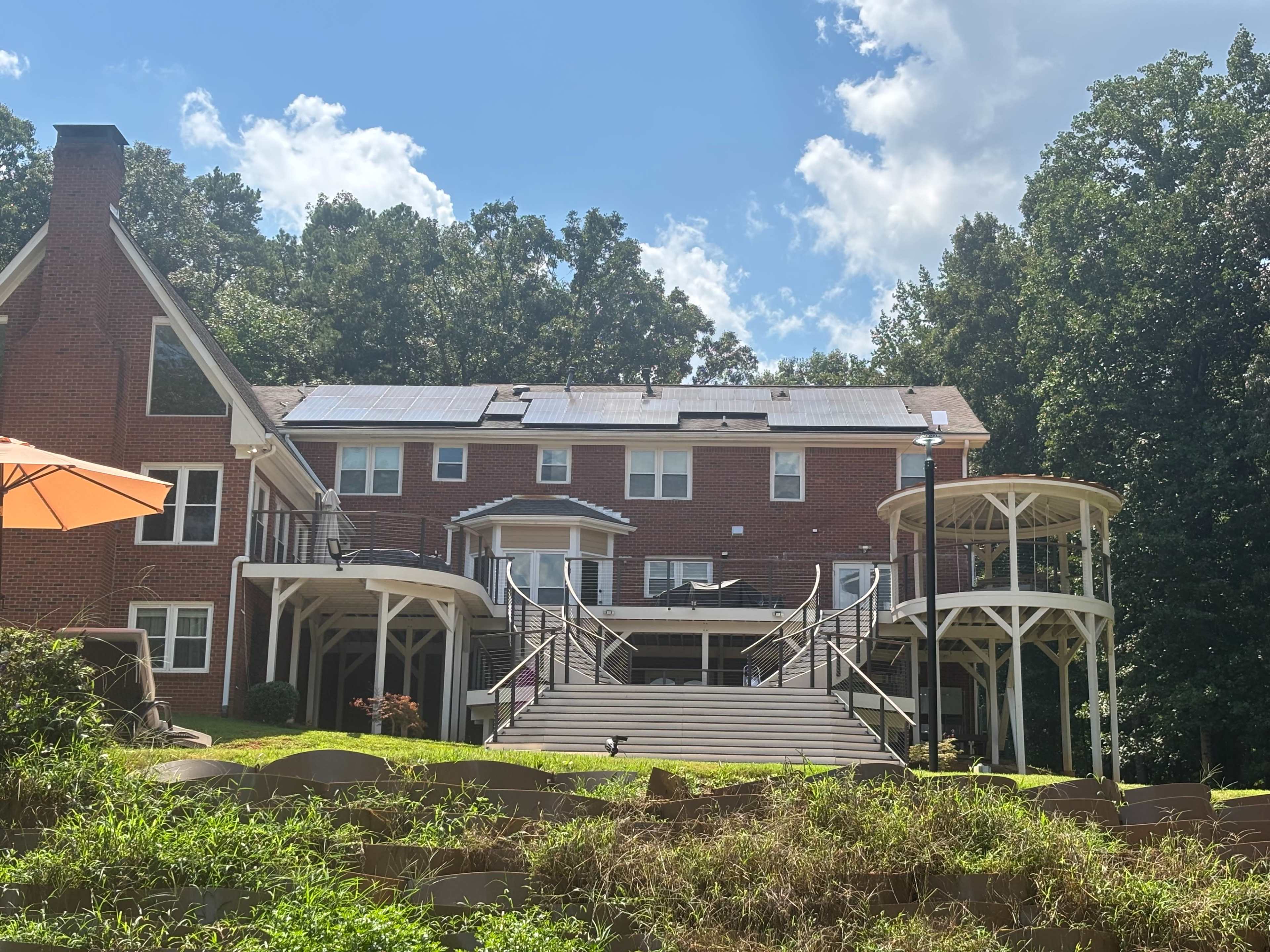 A large, red-brick house with solar panels on the roof and a two-level porch overlooks a grassy area surrounded by trees.