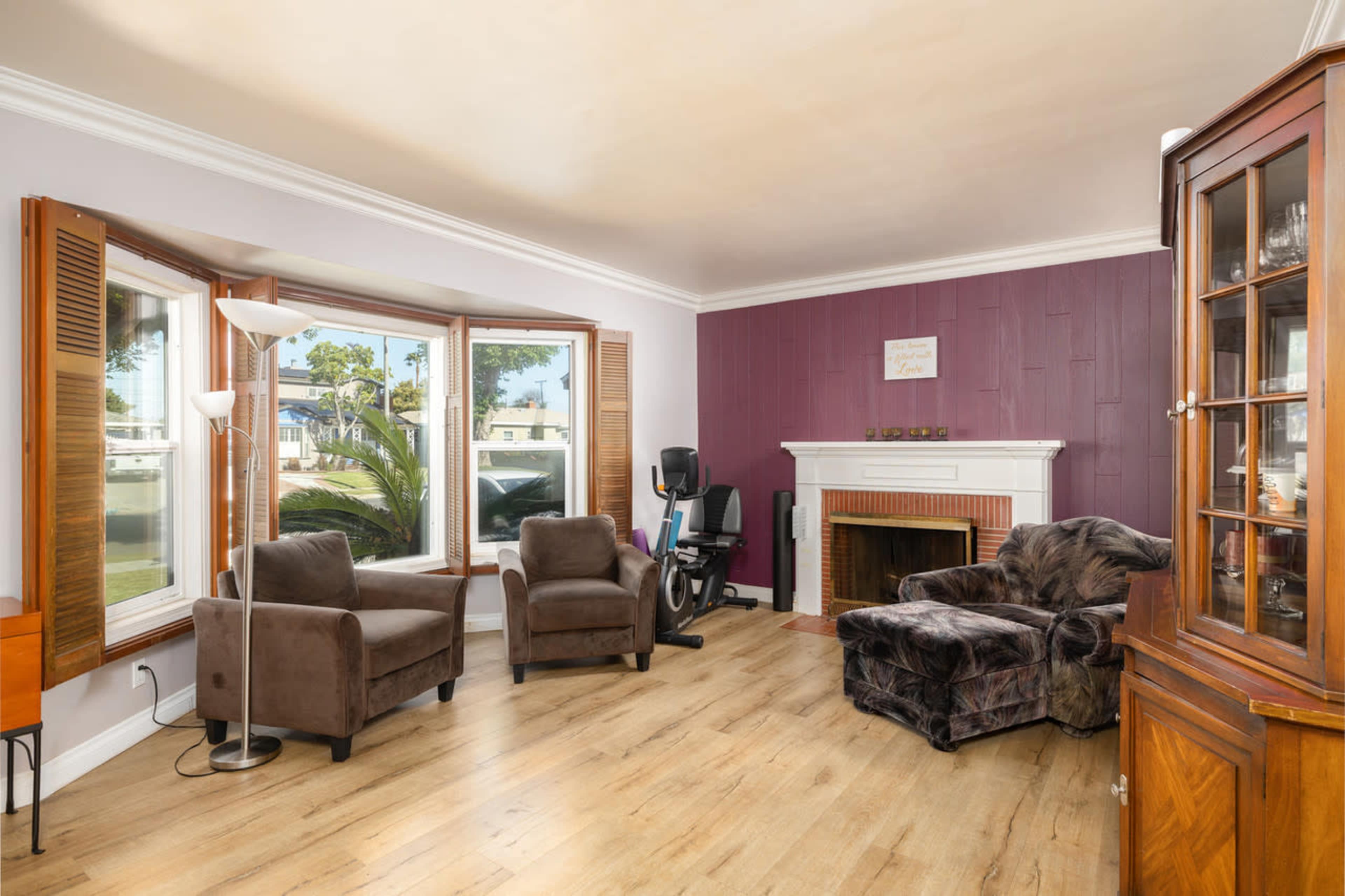 The image shows a cozy living room with two brown armchairs, a window nook, an exercise bike, and a fireplace, all set against a purple accent wall and wooden flooring.