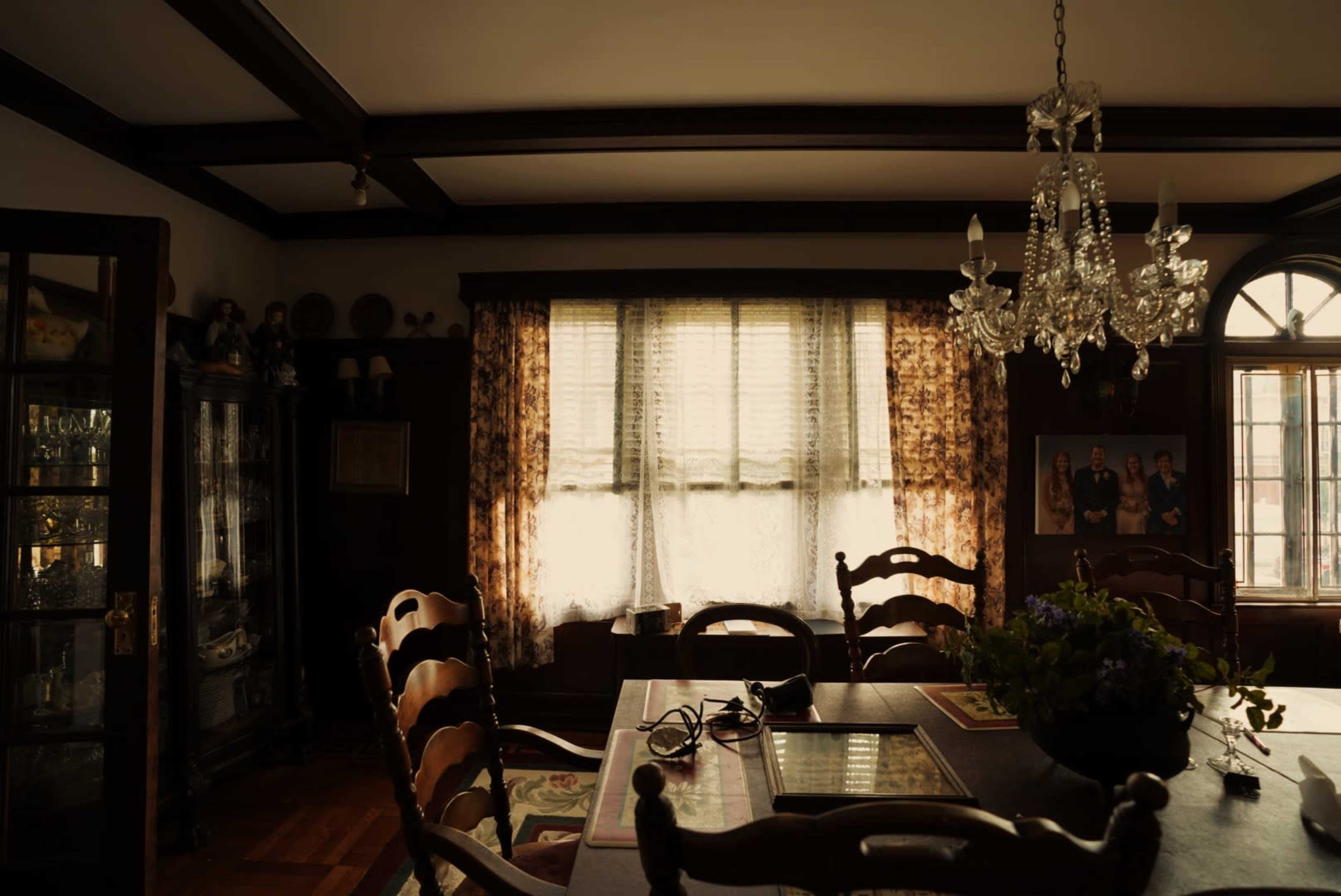 A dimly lit dining room with a large wooden table surrounded by seating, a chandelier overhead, and a window draped with sheer curtains.