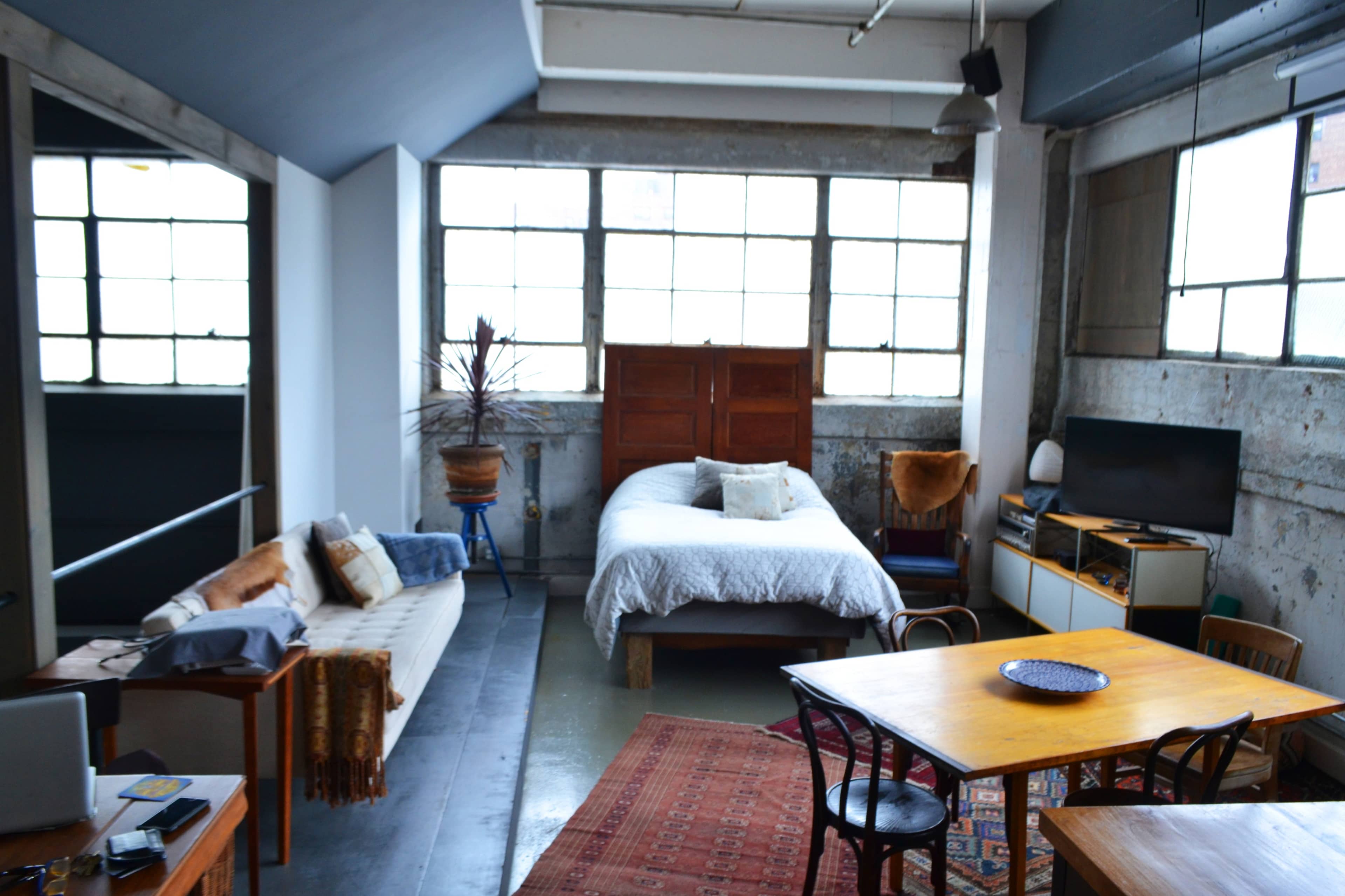 A spacious industrial-style bedroom with a bed, a dining table, and large windows illuminating the concrete walls.