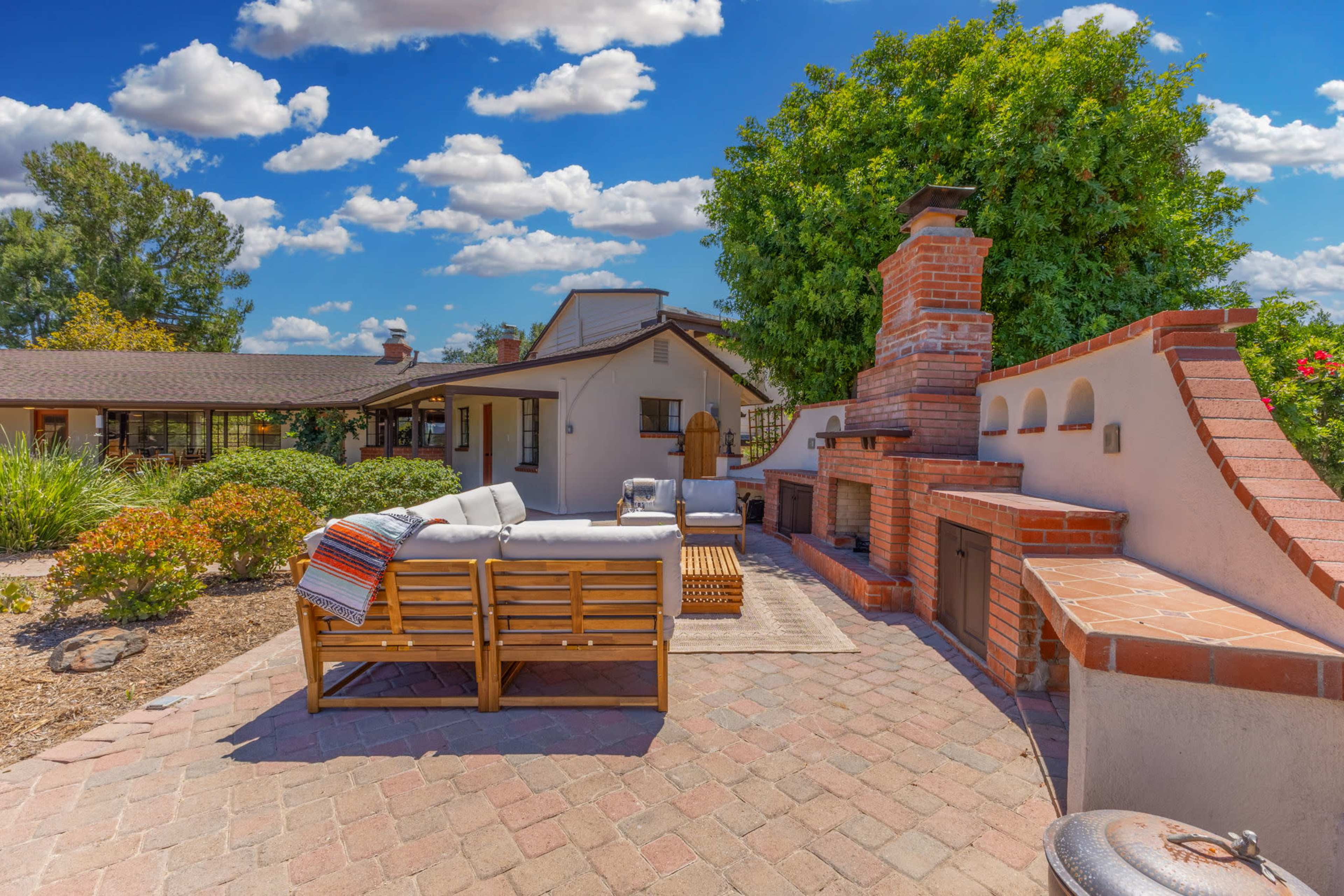 A patio area features a brick outdoor fireplace, wooden seating, and lush landscaping under a blue sky with clouds.
