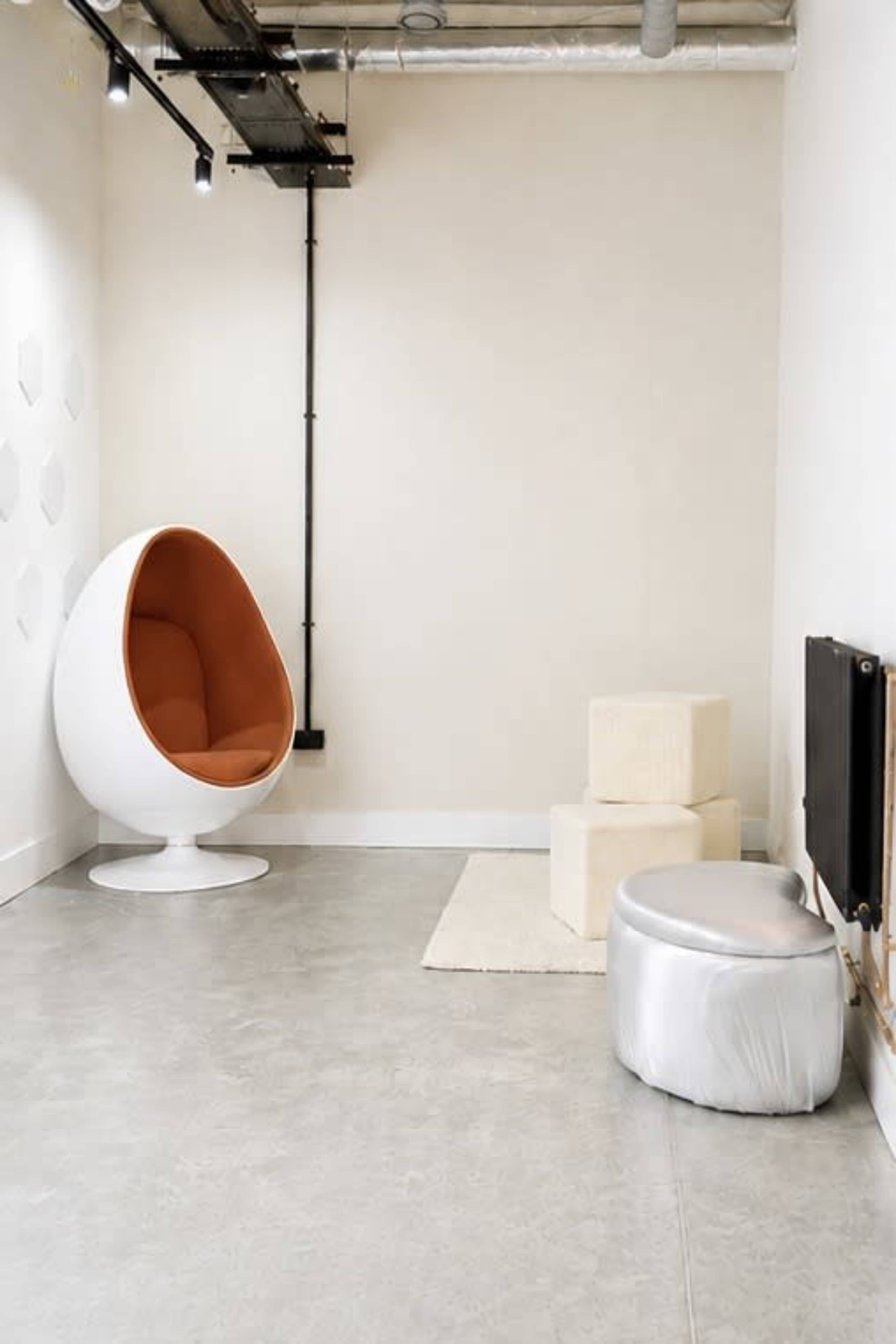 The image shows a modern, minimalistic interior featuring an orange and white egg-shaped chair, two light-colored cube stools, and a shiny silver ottoman on a gray floor.