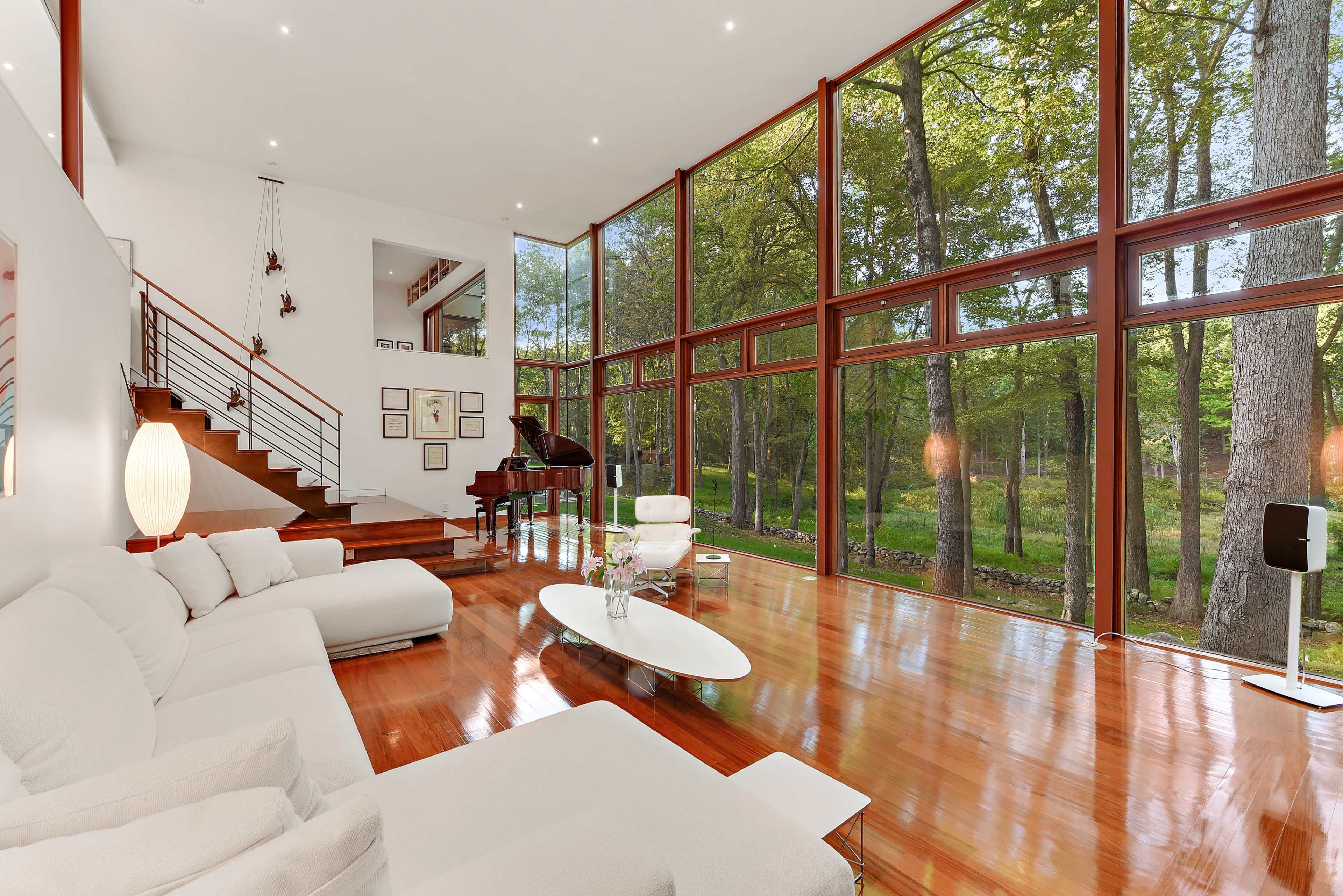A modern living room features large windows overlooking a wooded area, with a white sectional sofa and a piano on a polished wooden floor.