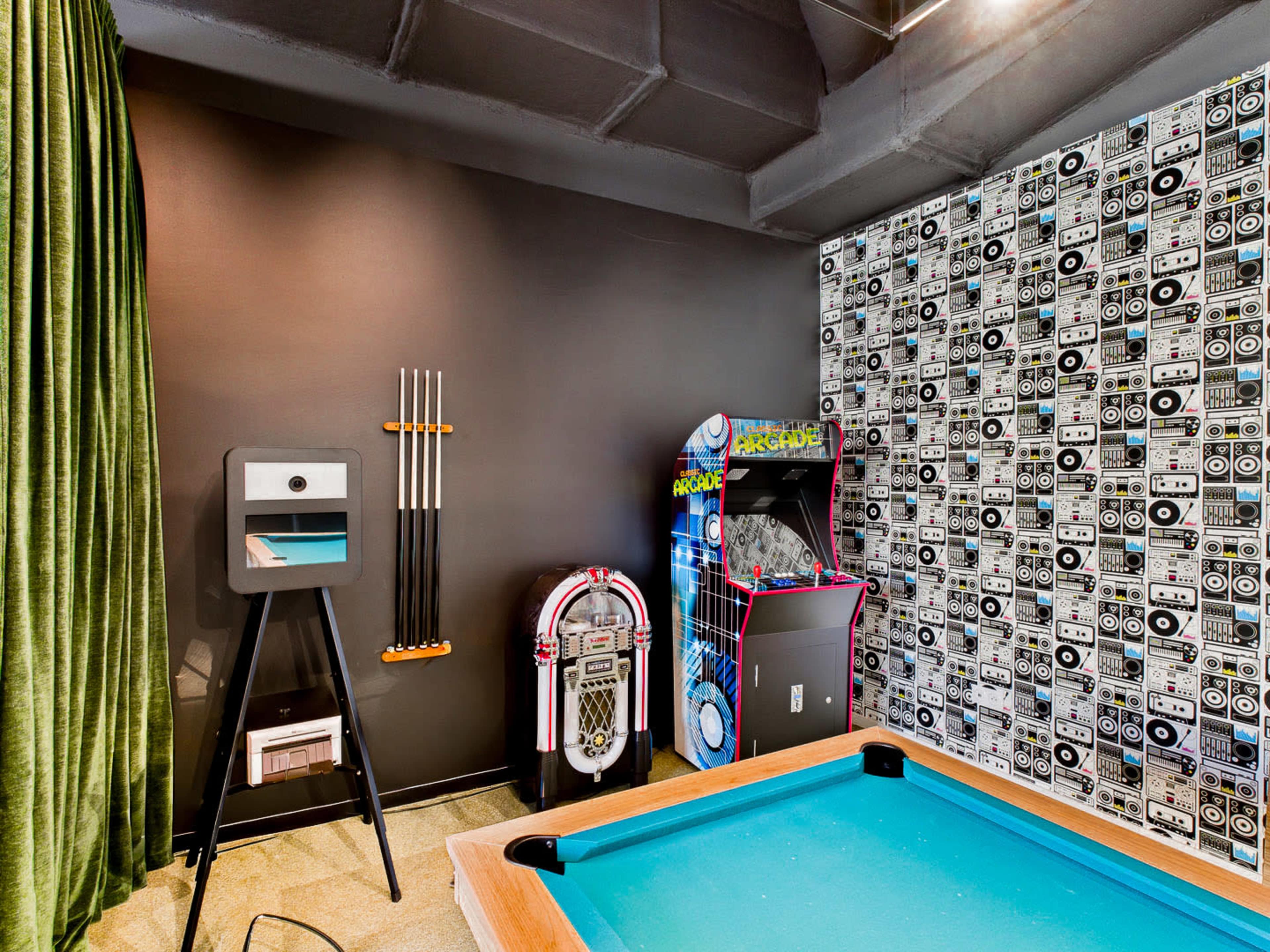 The image shows a recreational room with a pool table, a jukebox, an arcade game machine, and a green curtain against a patterned wall.
