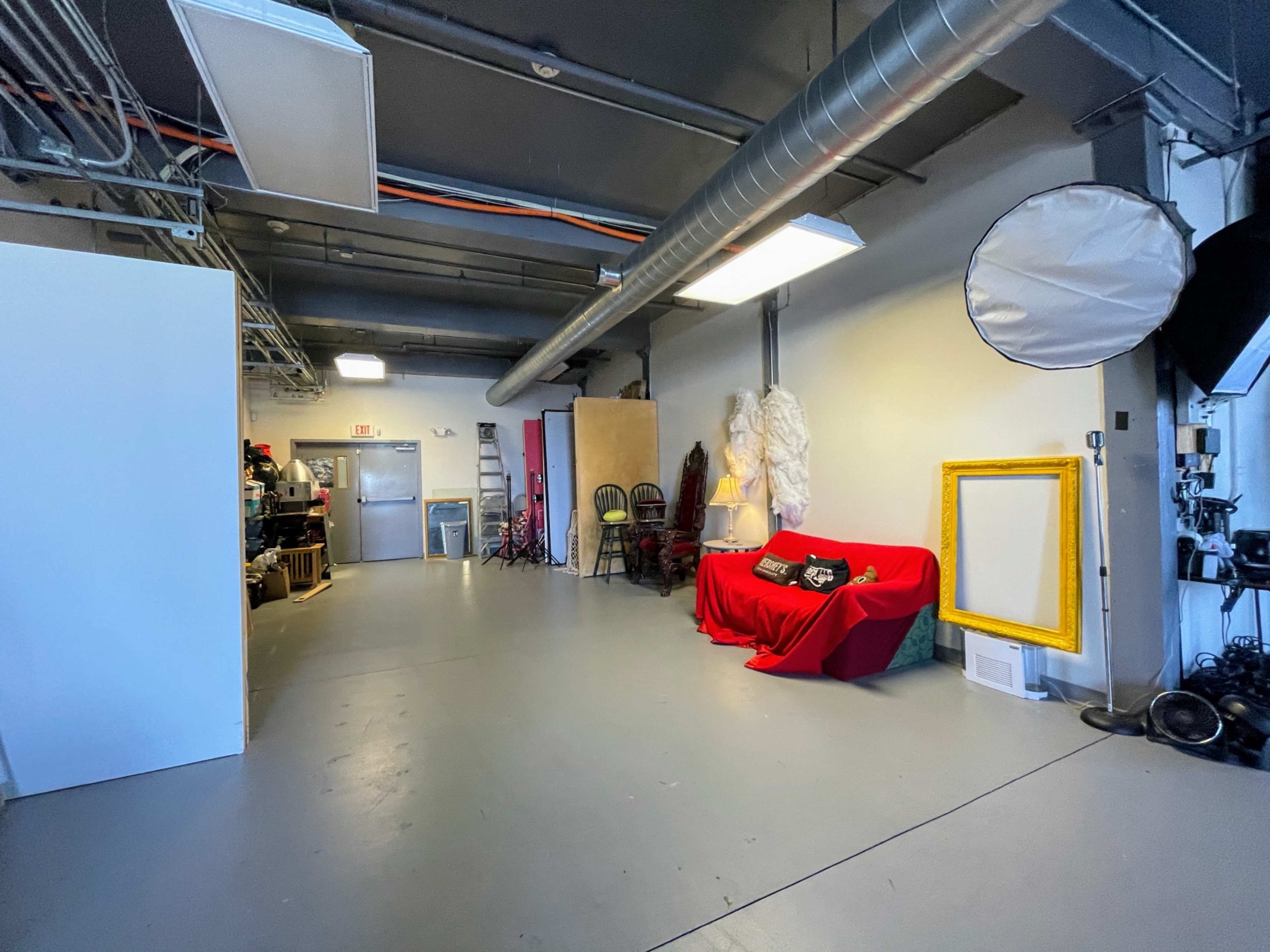 A large, industrial-style studio space featuring a red draped couch, a blank wall, a large frame, and various photography equipment in the background.