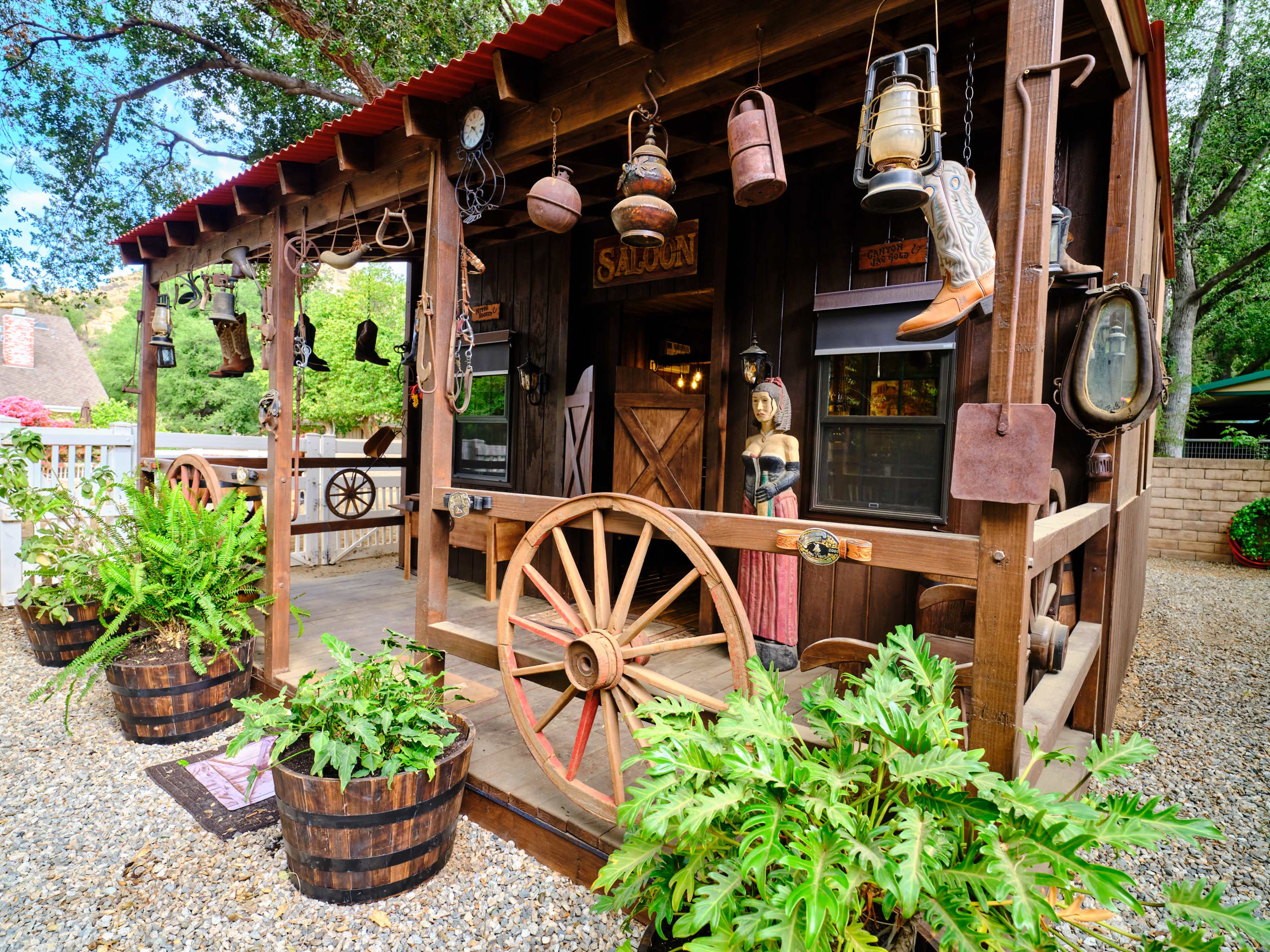 A rustic wooden structure features vintage decorations, including lanterns, cowboy boots, and a mannequin, surrounded by potted ferns and barrels.
