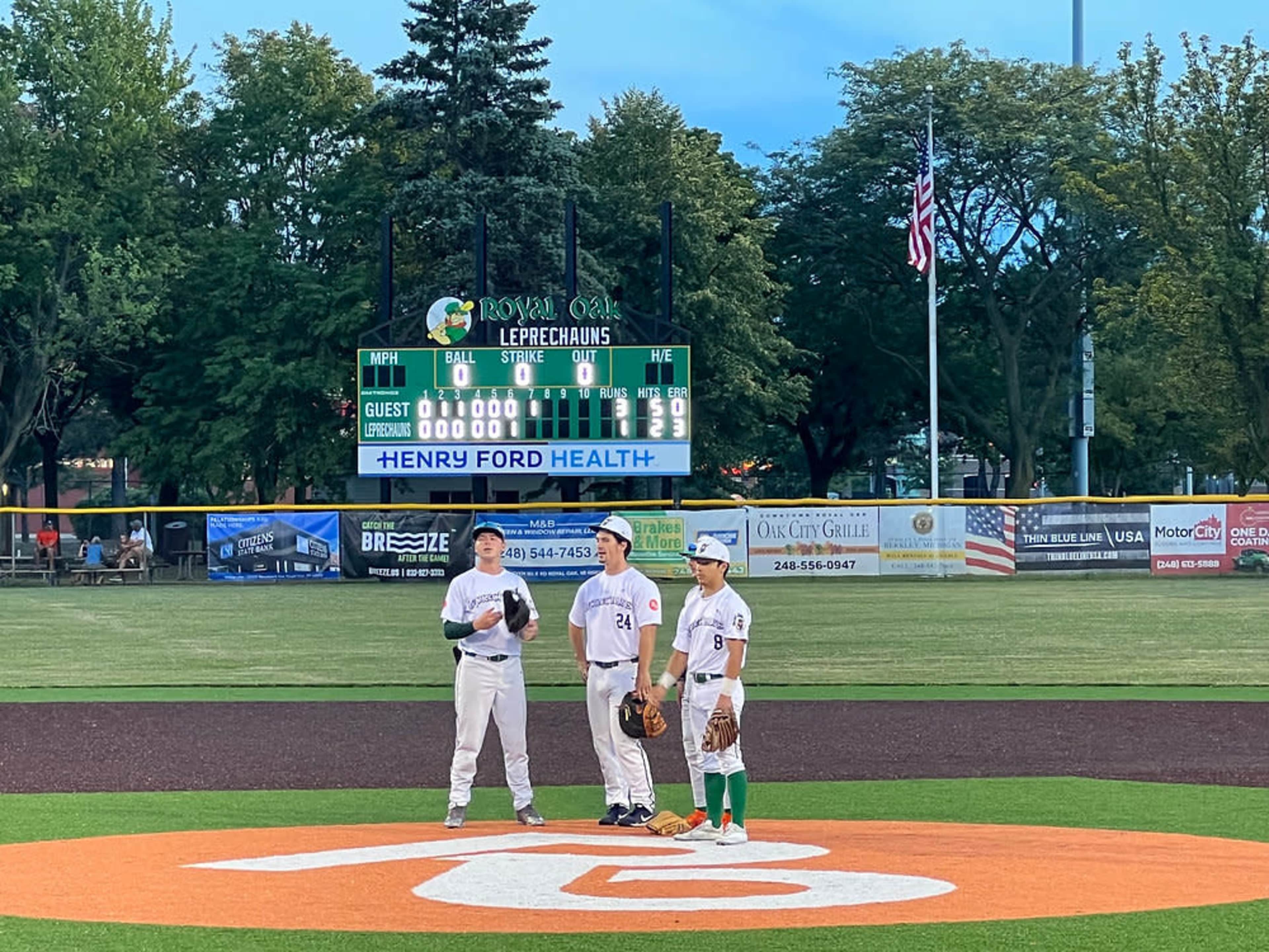 Three baseball players stand on the pitcher's mound during a game, with a scoreboard displaying the current game status in the background.