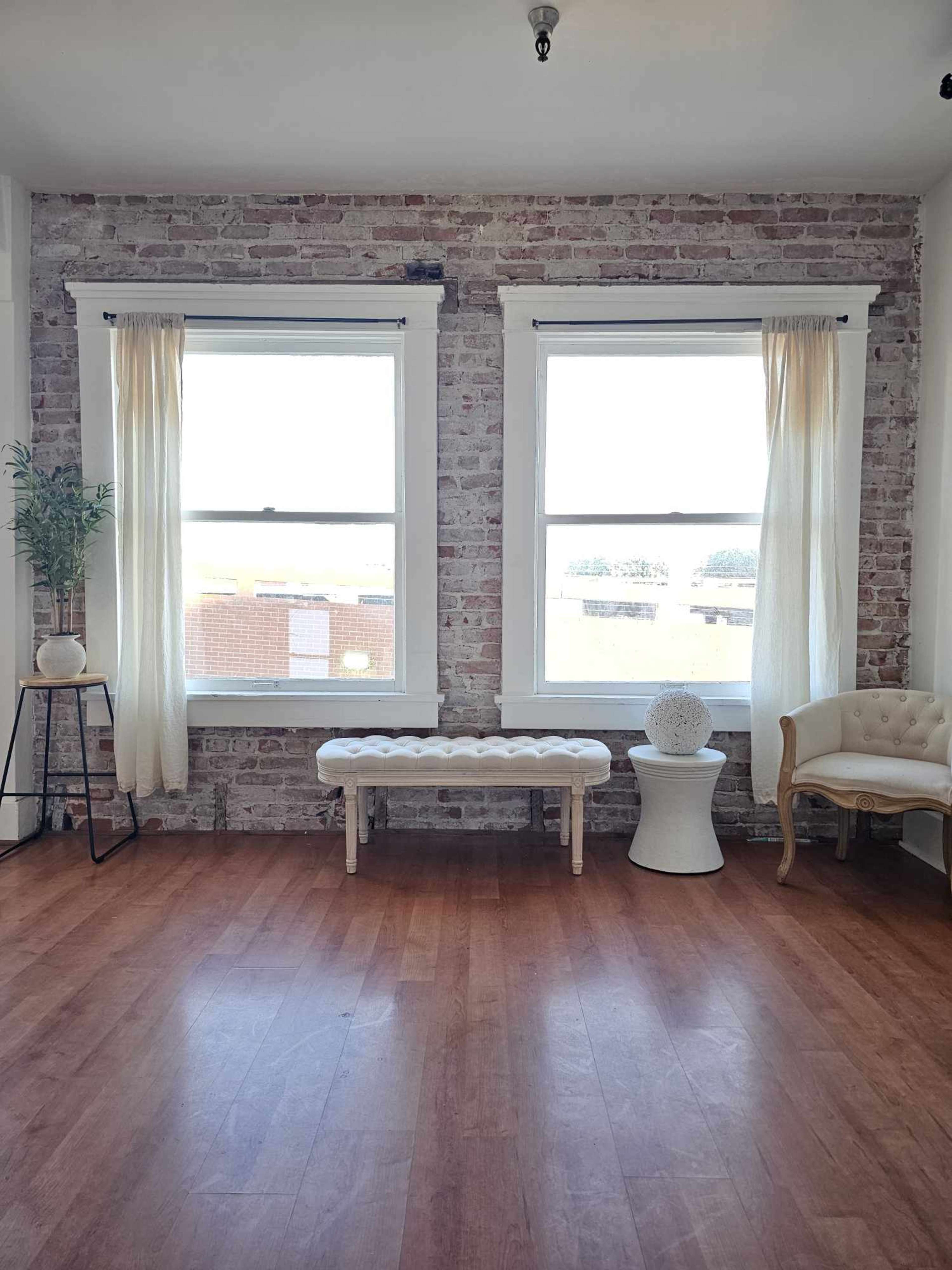 A well-lit room featuring two windows with sheer curtains, a wooden floor, and various furniture pieces, including a tufted bench and a decorative stool.