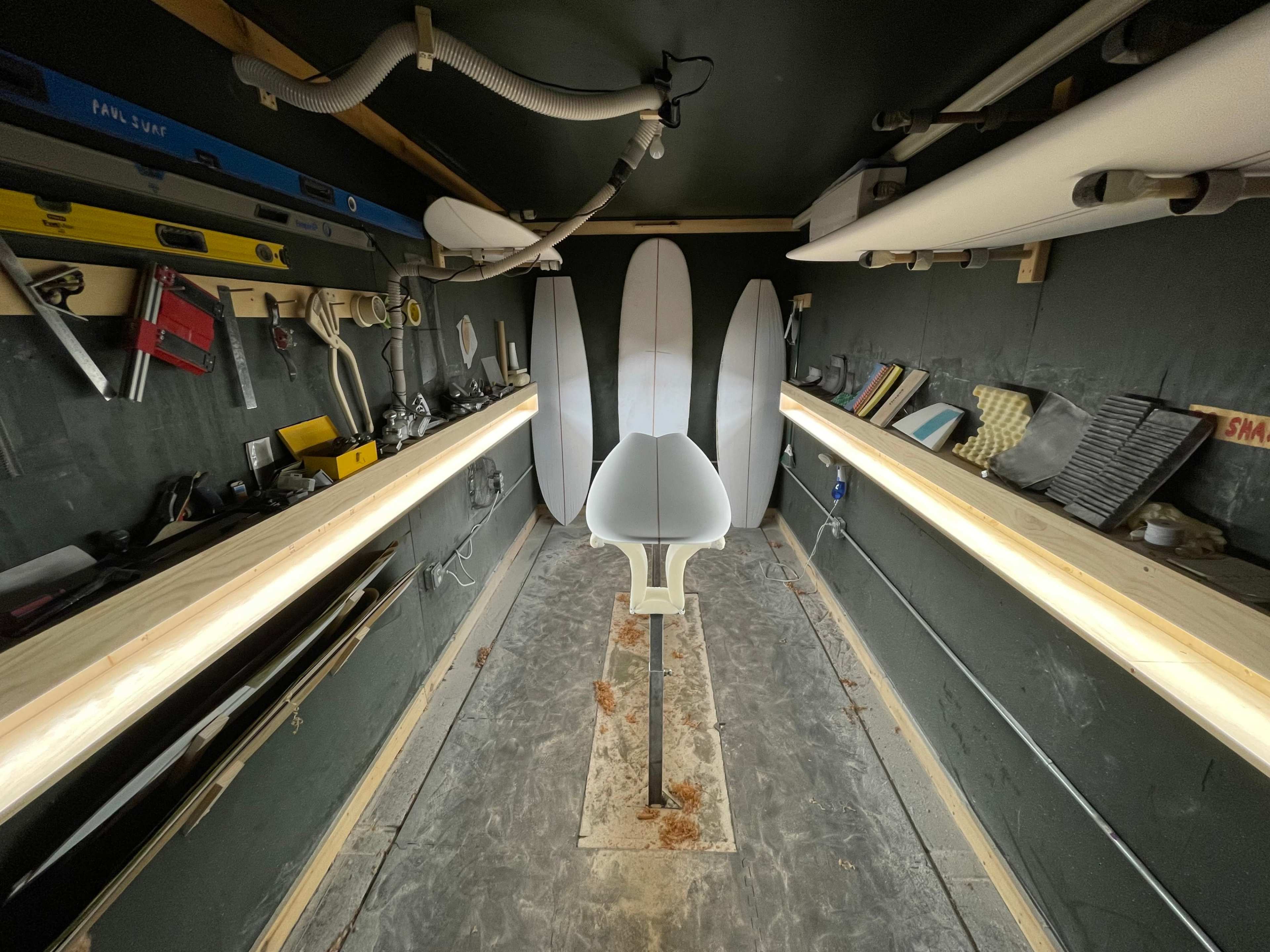The image shows a workshop with surfboard shaping tools, including several surfboard blanks, illuminated by linear lights along the walls.