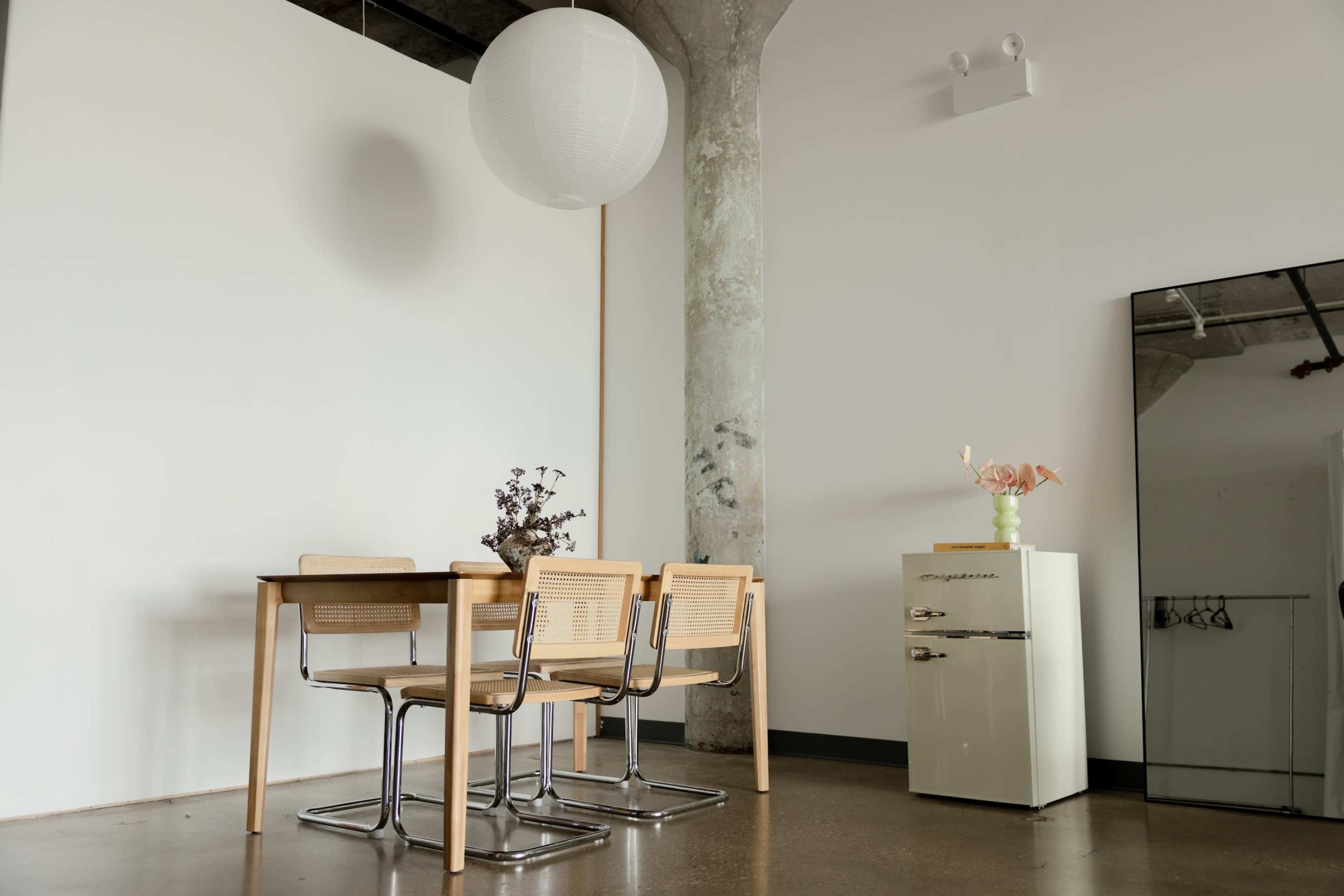 The image shows a minimalist dining area with a wooden table and three chairs, a small flower vase, a vintage cream-colored fridge, and a large mirror against a plain wall.