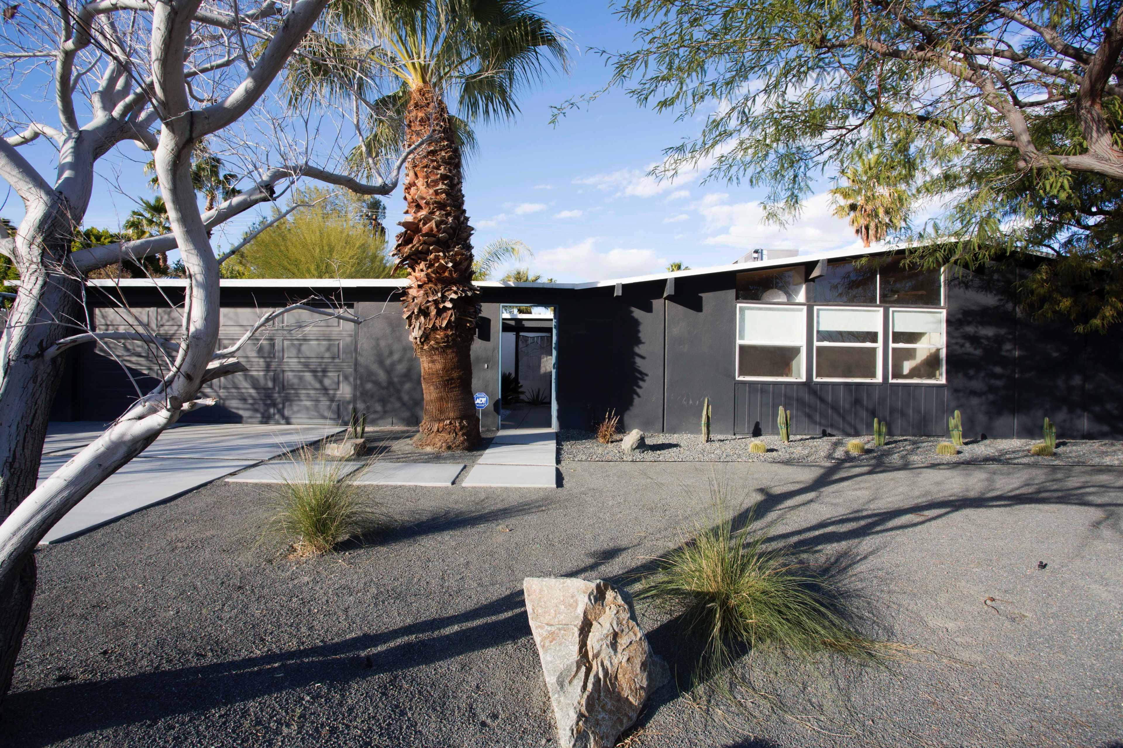 A modern black house with a gravel yard, surrounded by palm and desert plants, features a concrete driveway and large front windows.