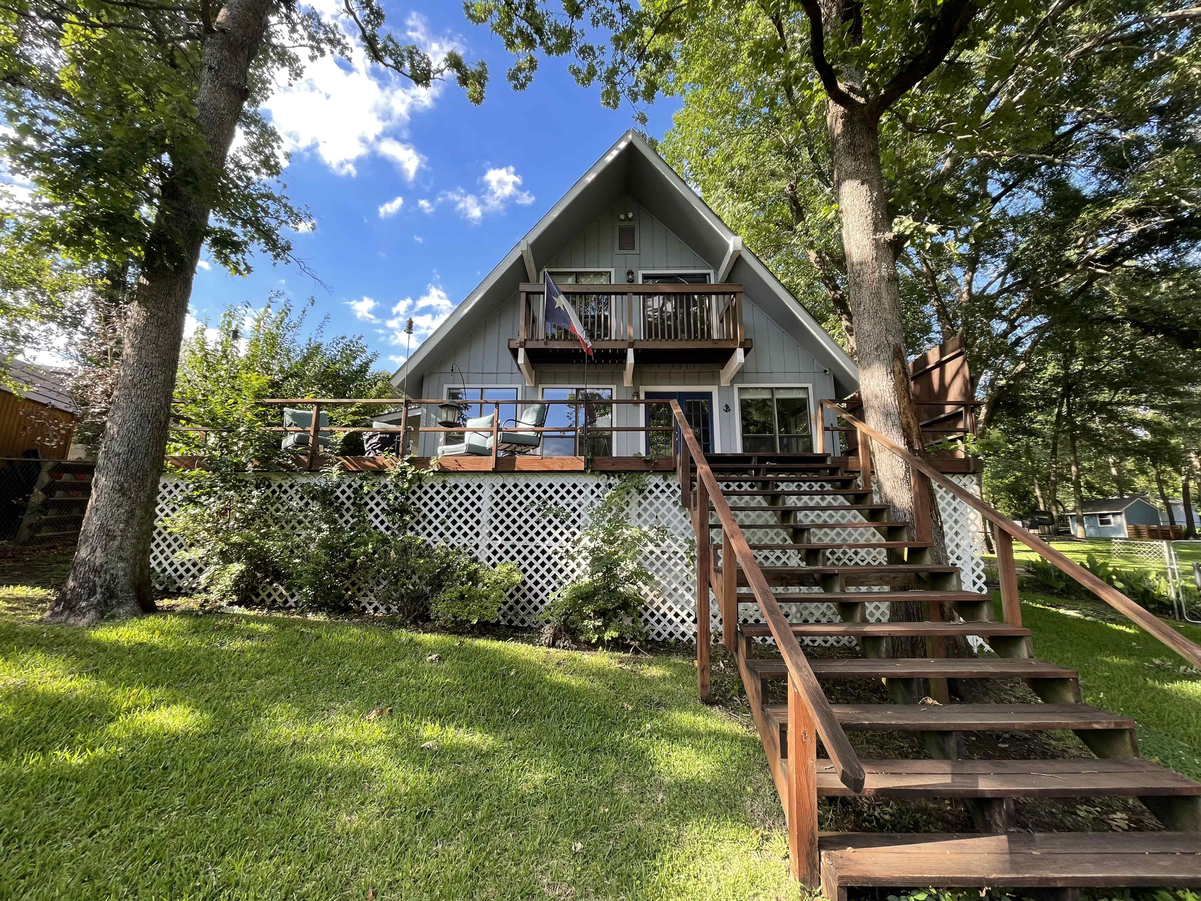 A two-story A-frame cabin with a wooden deck and stairs, surrounded by trees and lawn, is set against a blue sky with scattered clouds.