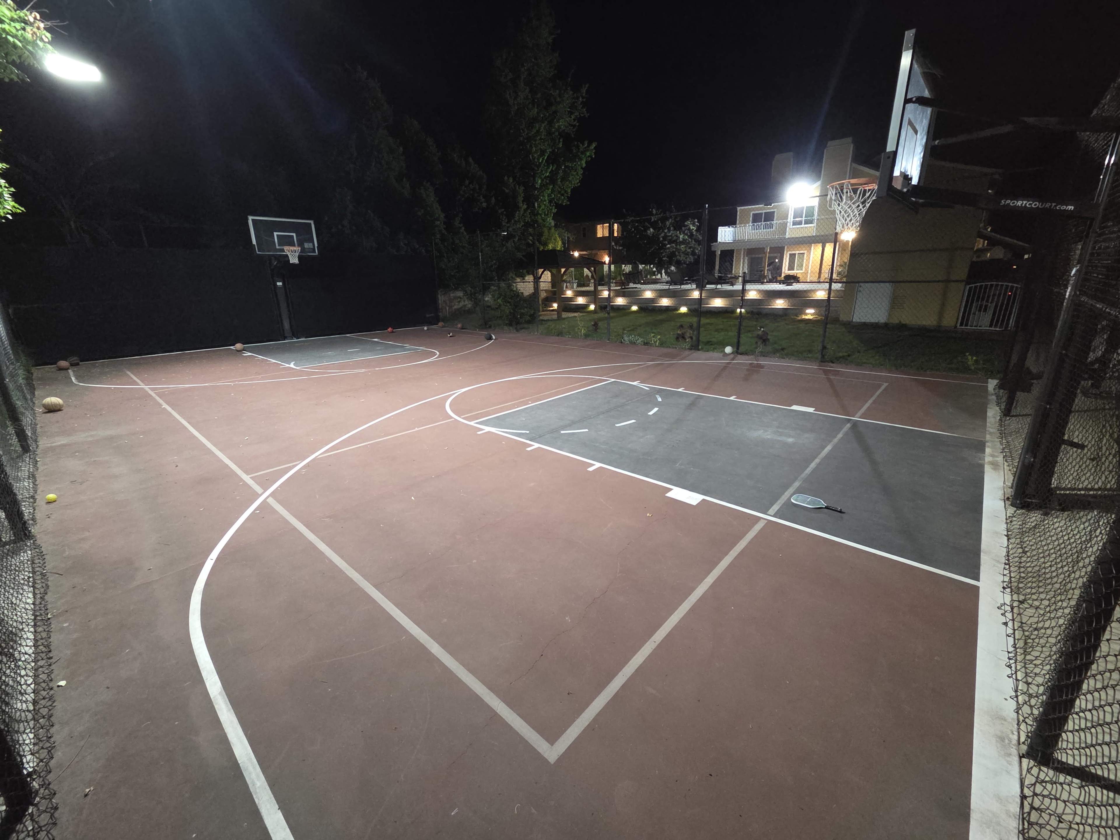A well-lit outdoor basketball court features two playing areas, surrounded by nets and trees, under a night sky.