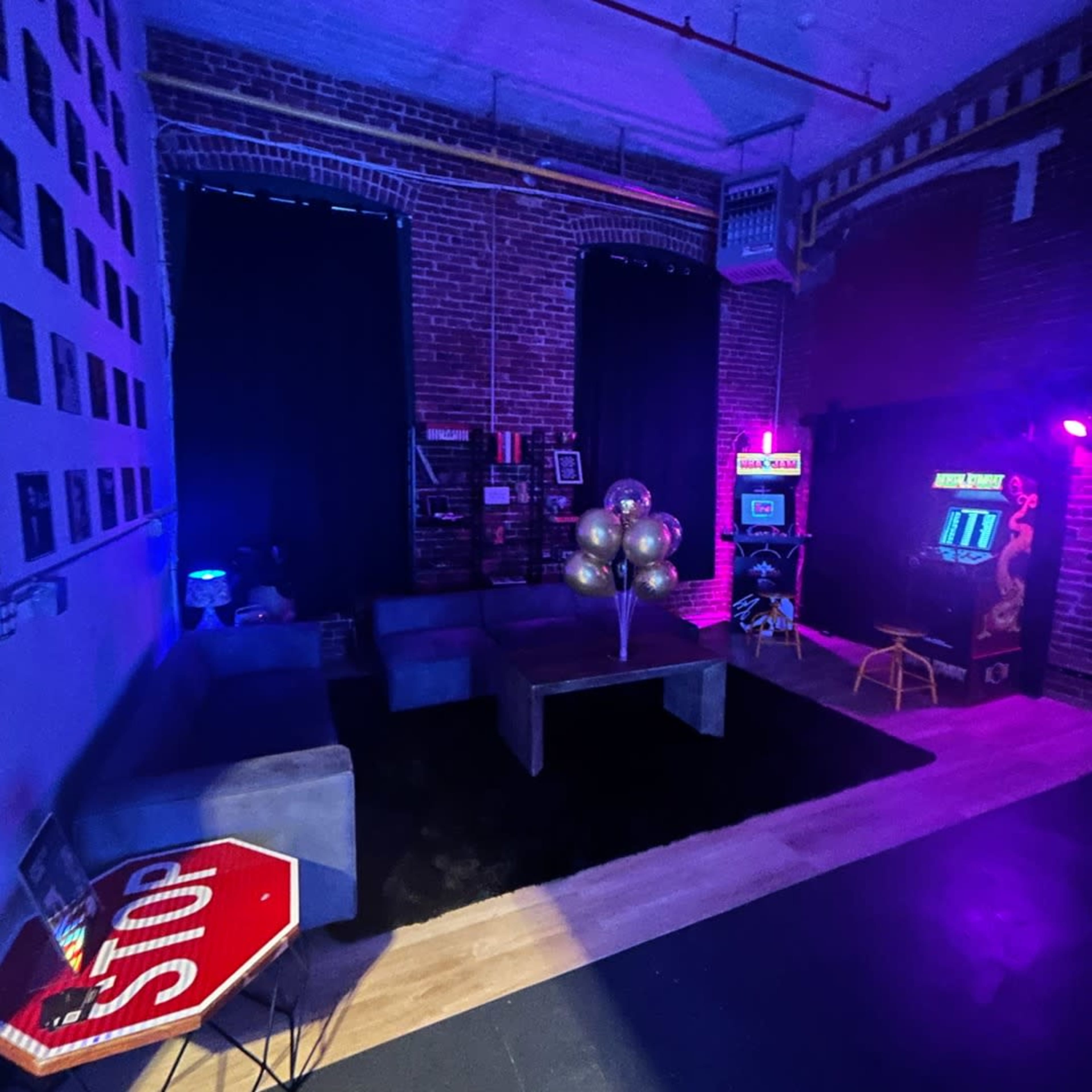 A dimly-lit lounge area with brick walls, featuring a black rug, comfortable seating, a table with a decorative balloon arrangement, and an arcade machine in the background.