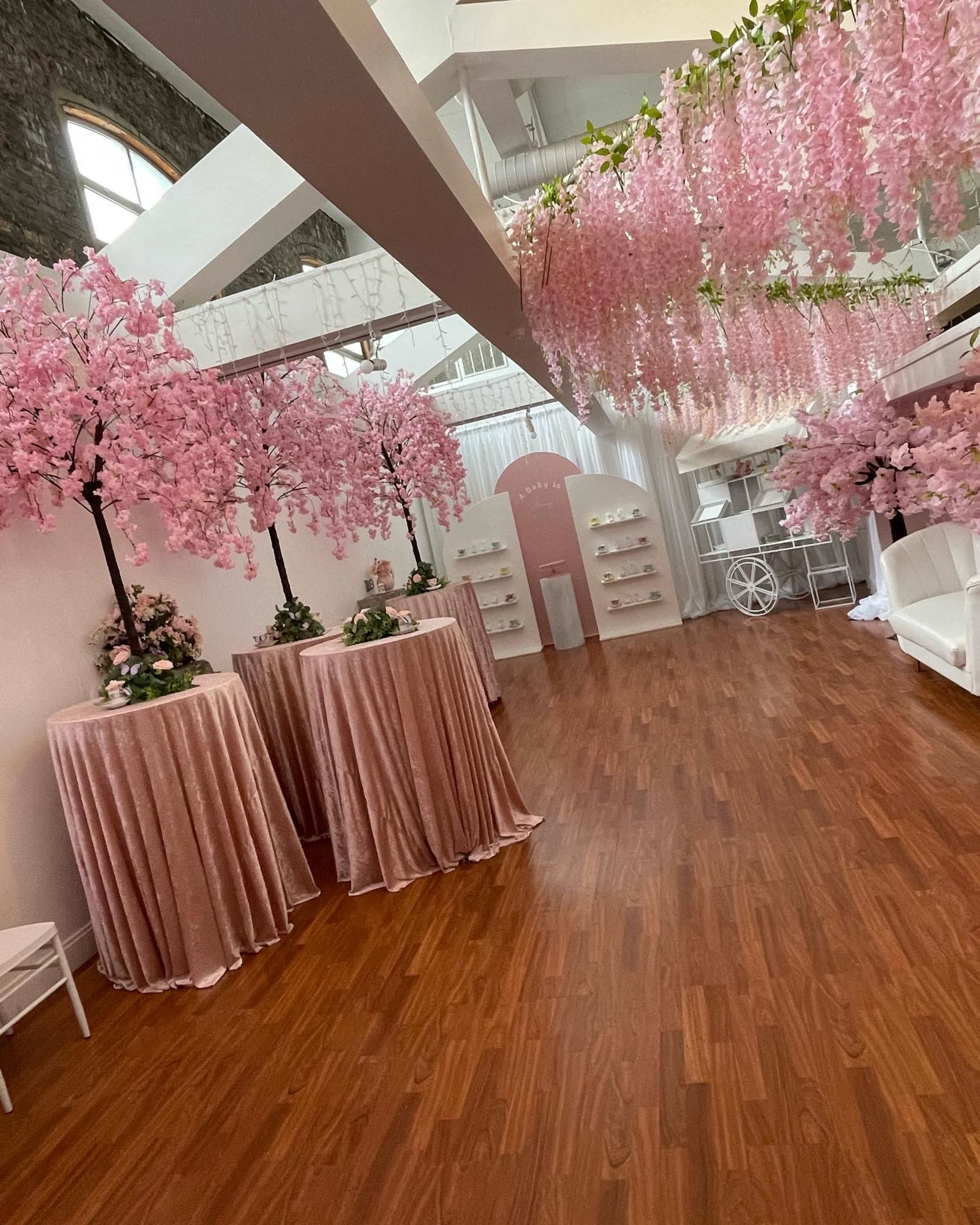 The image shows a spacious room decorated with pink wisteria trees, velvet-covered tables, and white decor elements, creating an elegant atmosphere.