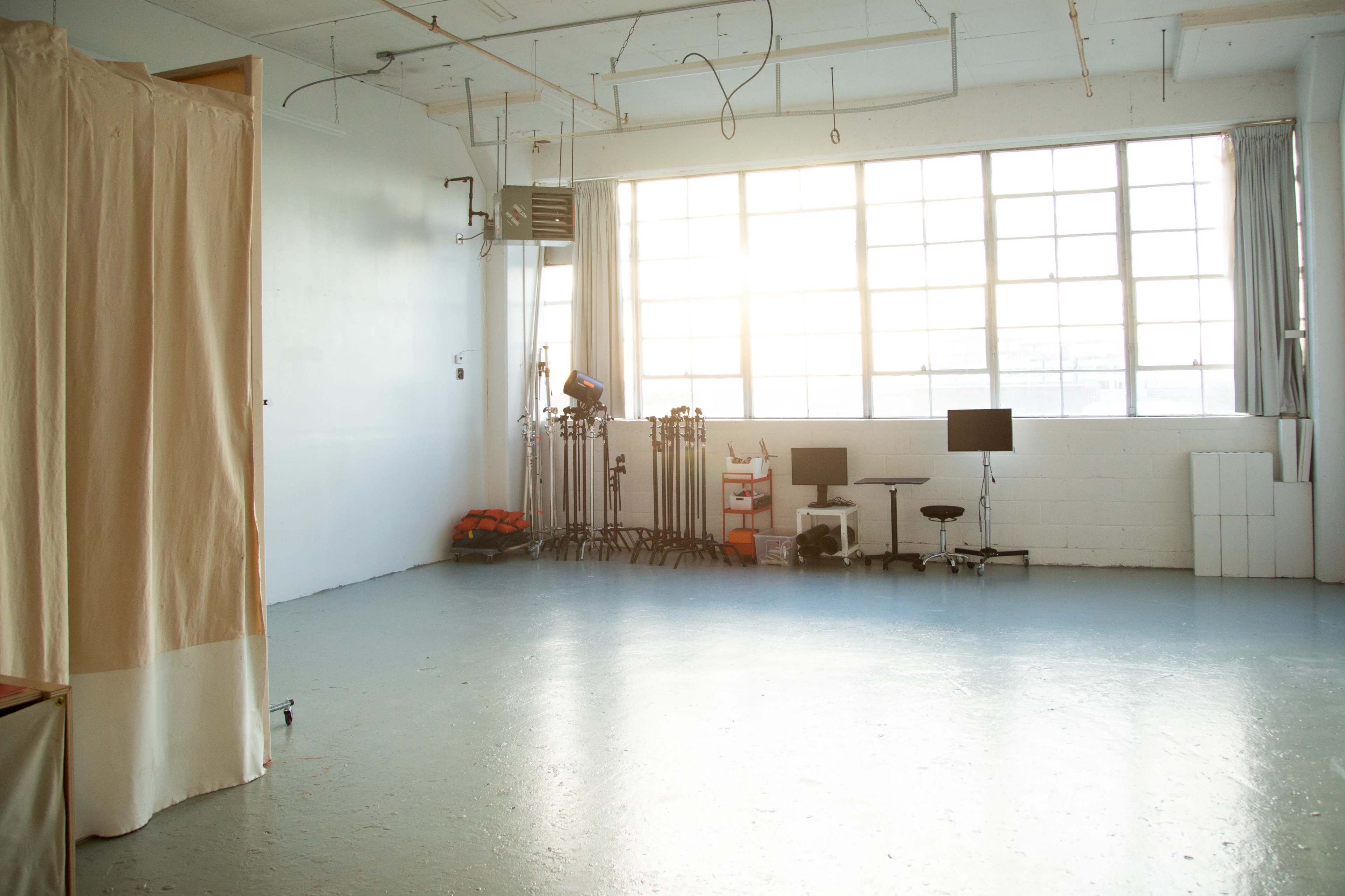 An empty studio space with large windows, beige curtains on one side, and equipment set up including two computer monitors and stands.