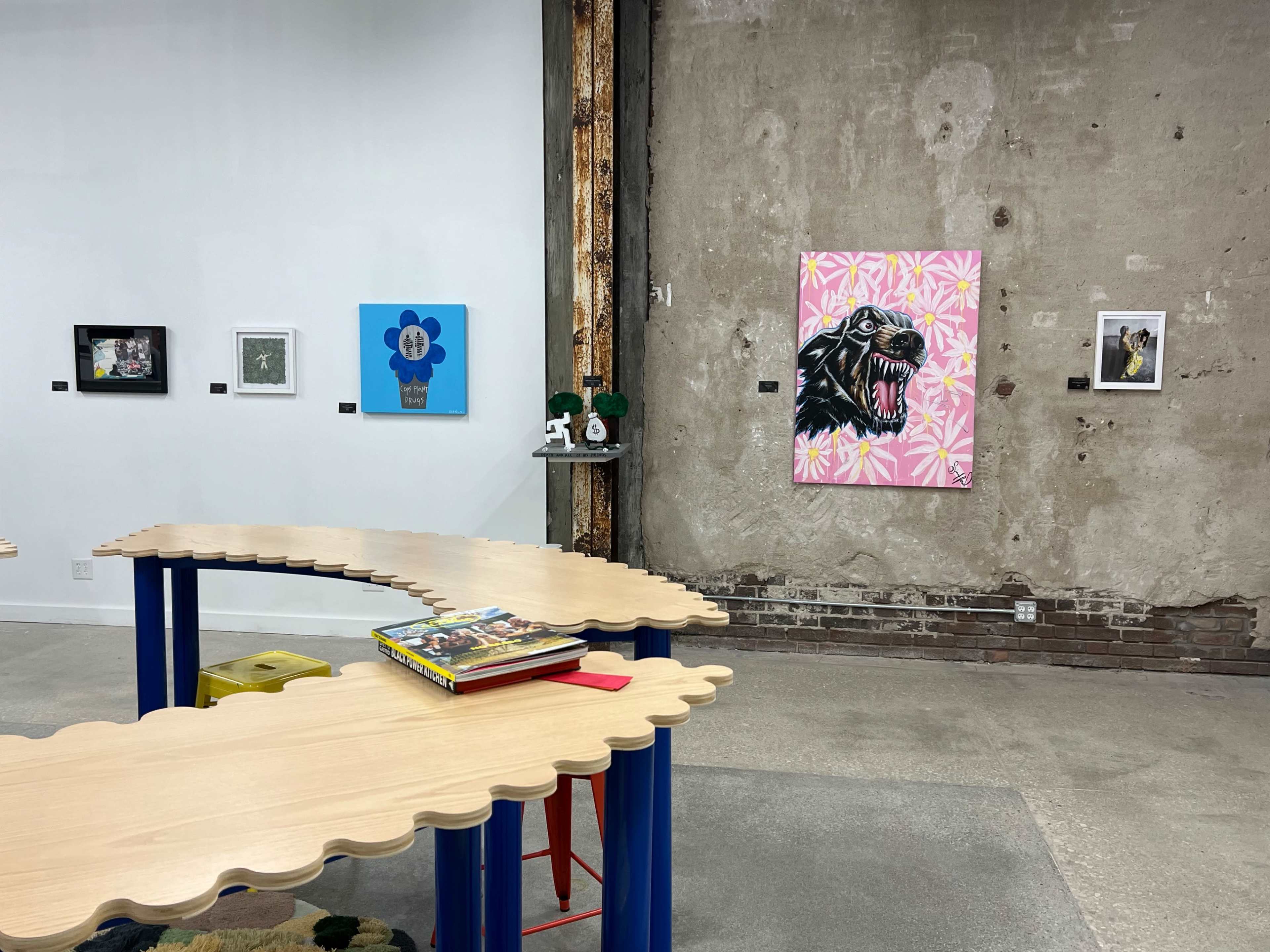 The image shows an art gallery with several framed artworks displayed on a gray wall, a unique wavy wooden table in the foreground, and various decorative items on the table.