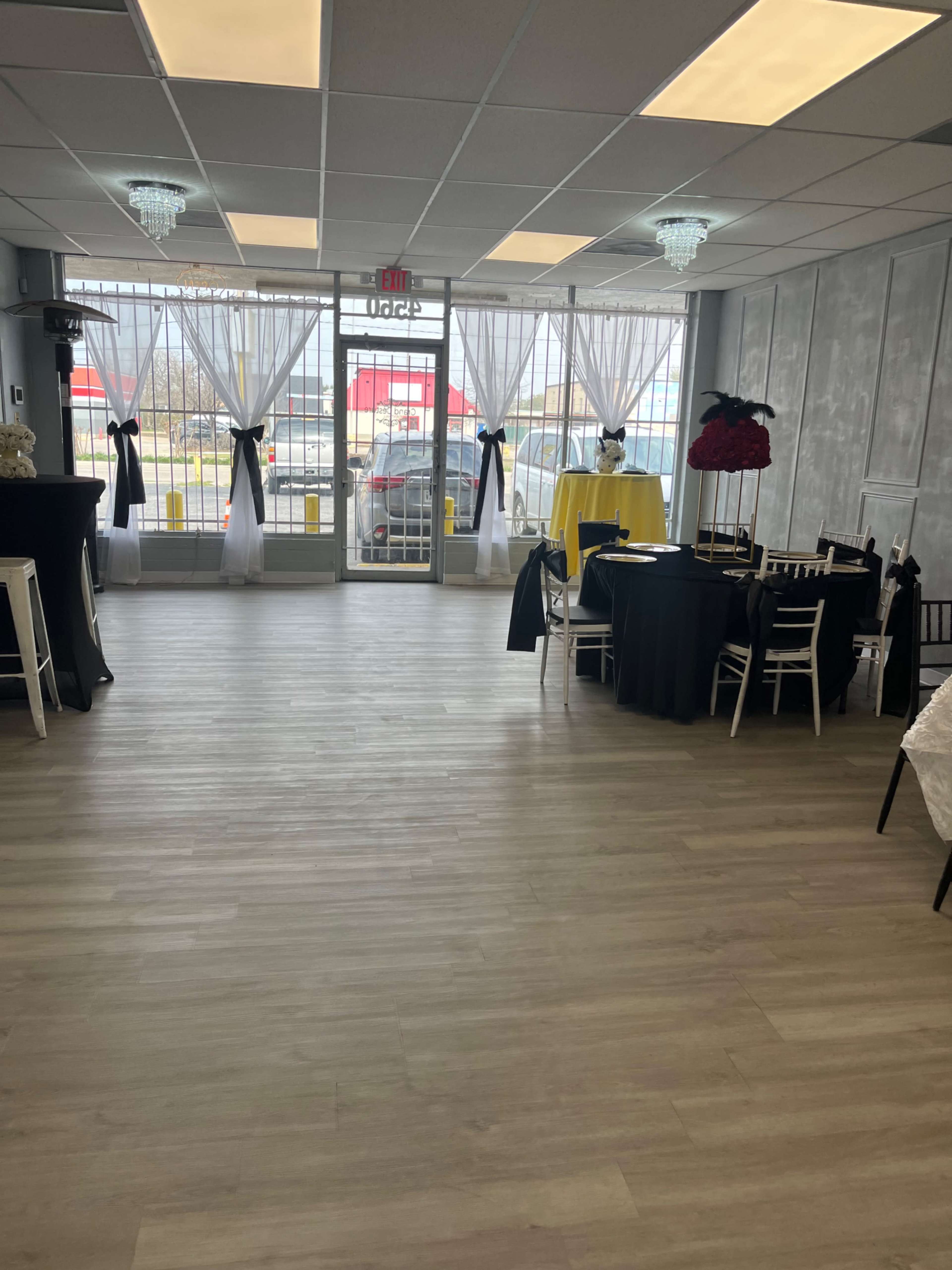 The interior of a decorated event space features tables with various tablecloths and chairs, large windows with sheer curtains, and decorative light fixtures.
