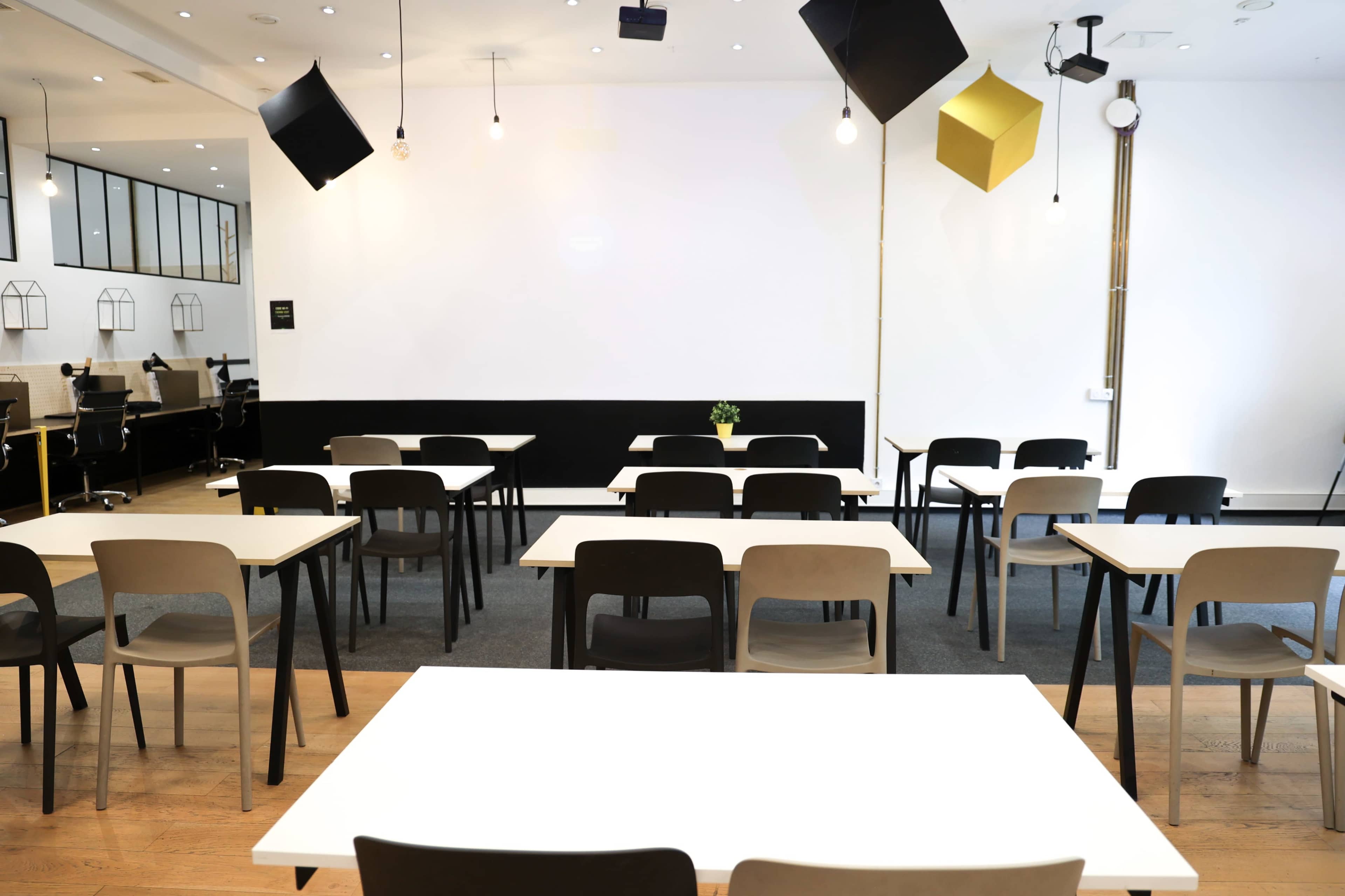 The image depicts a modern, empty classroom or meeting space with rows of tables and chairs, featuring a minimalist design and a white wall backdrop.