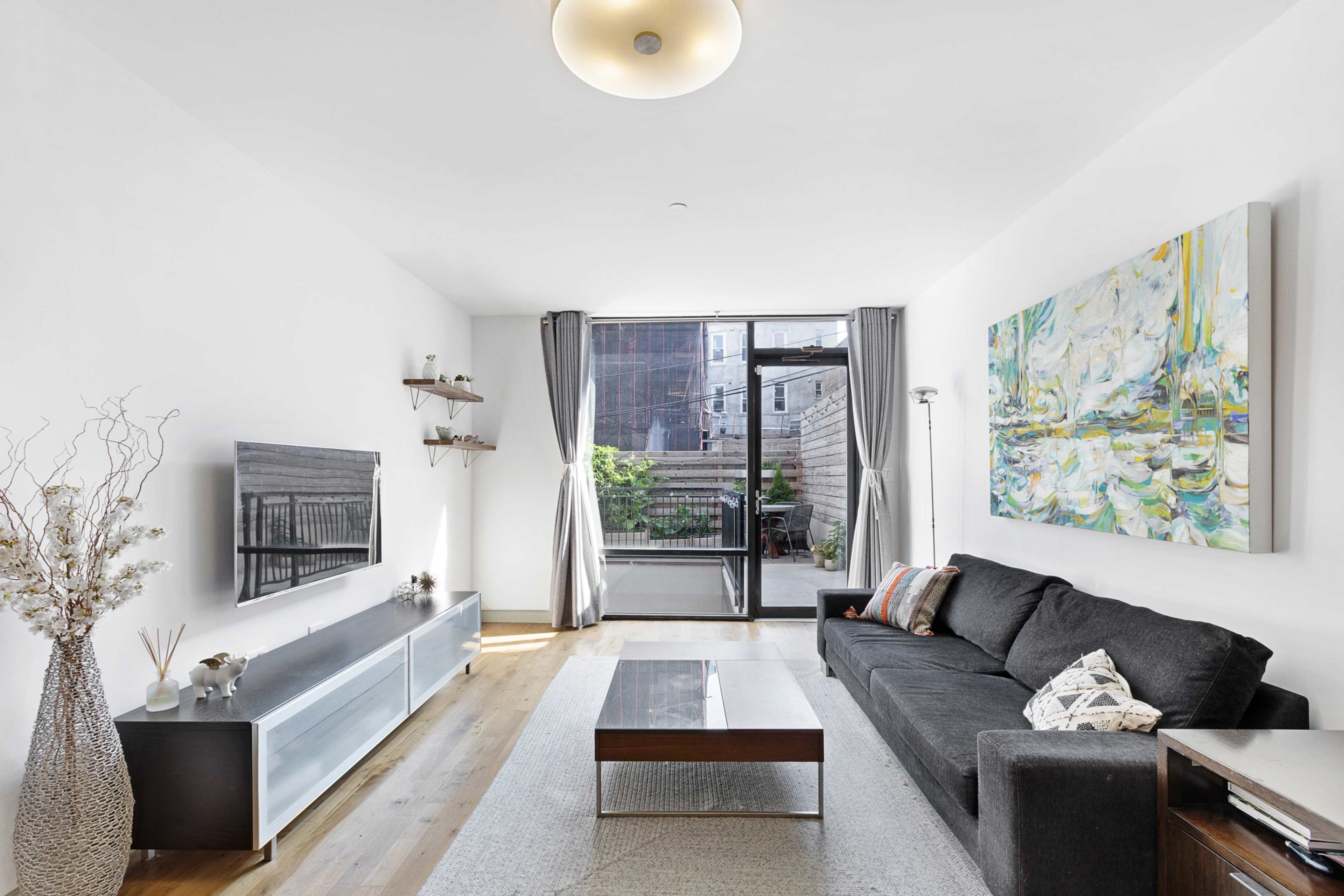 A modern living room features a dark sofa, a large abstract painting on the wall, and a glass door leading to a patio.
