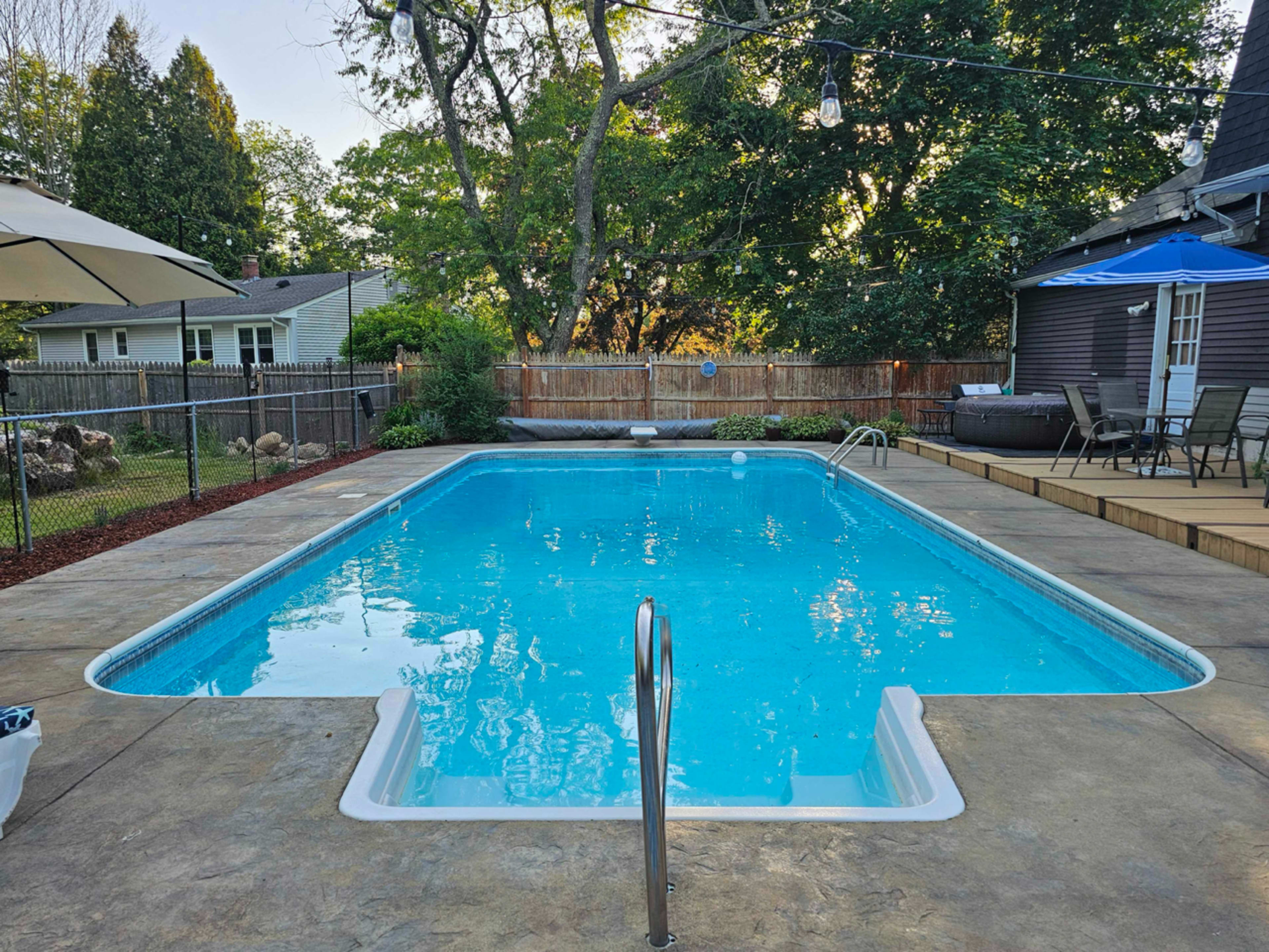 A clear blue swimming pool surrounded by a wooden deck and a fenced backyard with trees and a house in the background.