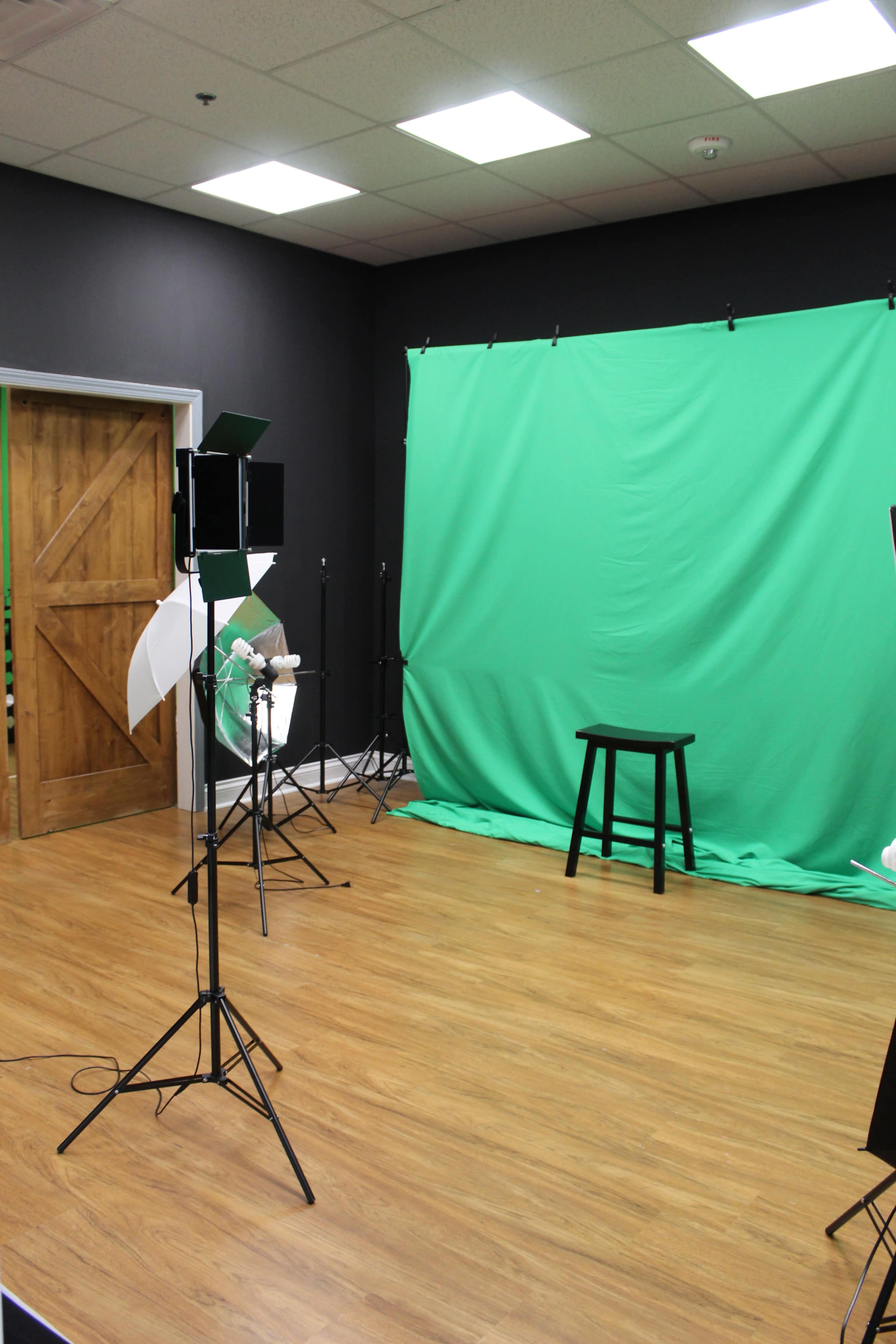 The image shows a photography studio with a green backdrop, a stool, and lighting equipment arranged in a spacious room.